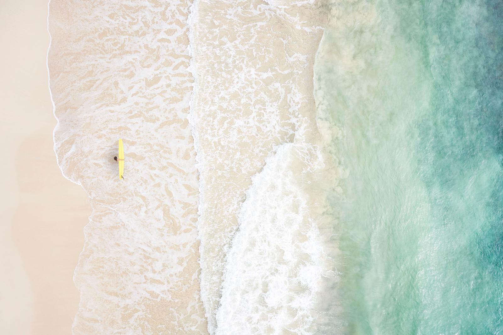 Un surfista solitario con una tavola gialla cammina sulla riva alla ricerca dell'onda perfetta - Photograph di Drew Doggett