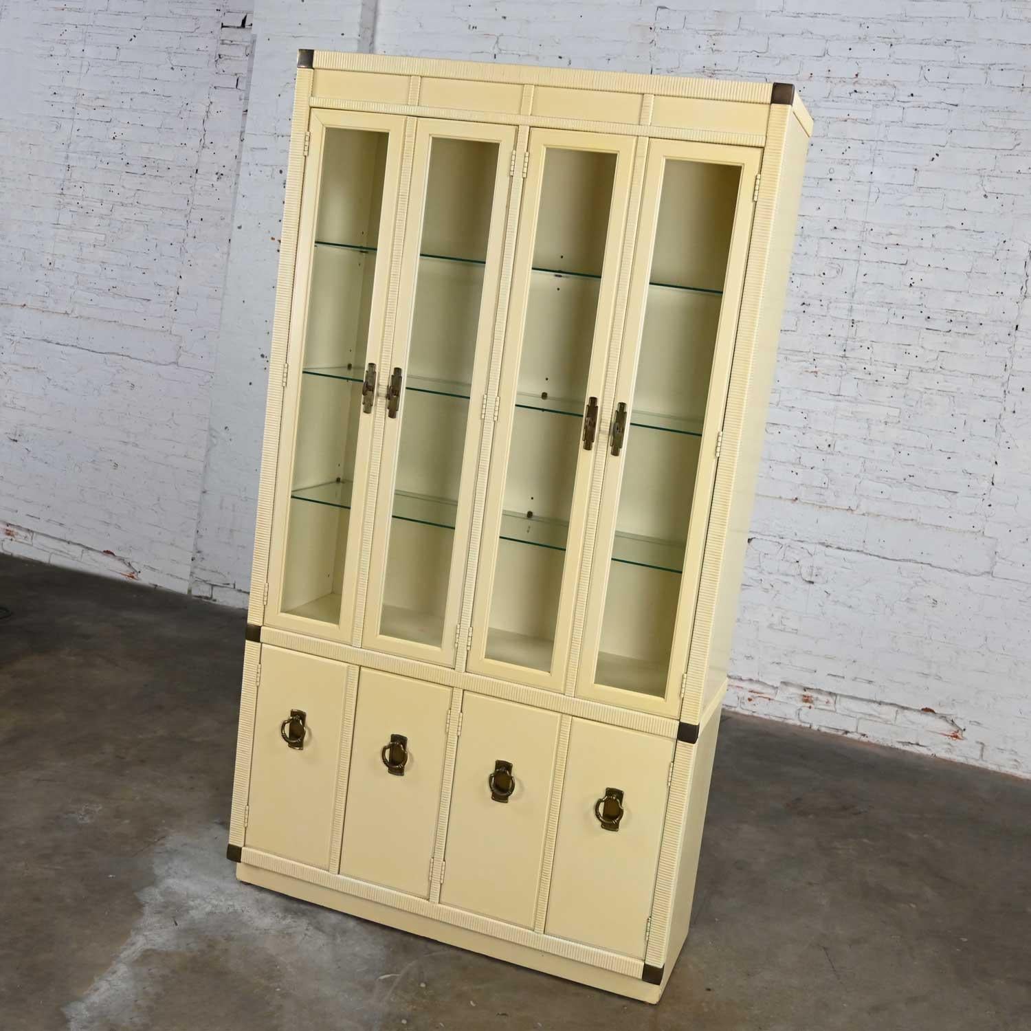 Lovely vintage Chinoiserie or Campaign style off-white lighted display cabinet with faux wrapped rattan edges by Drexel. Beautiful condition, keeping in mind that this is vintage and not new so will have signs of use and wear. There are a few dings