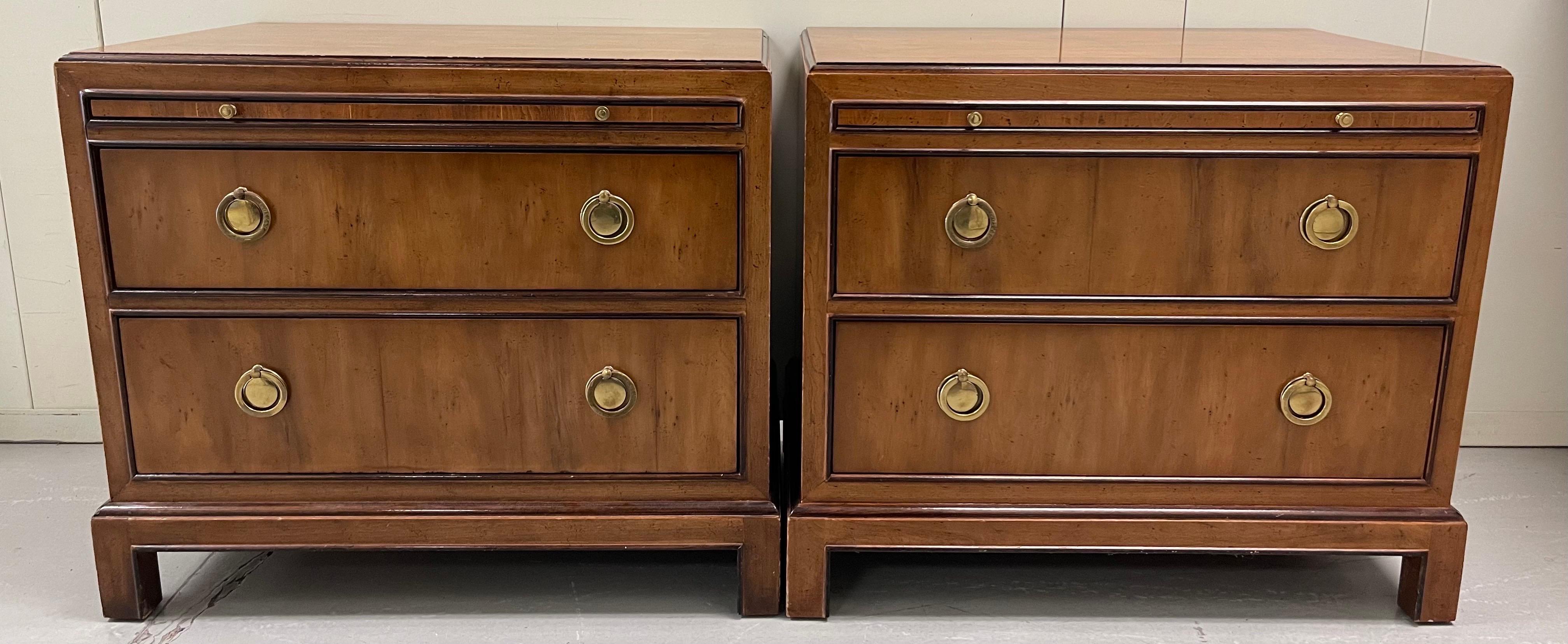 Pair of Drexel Heritage campaign style Burlwood nightstands. Original stained wood finish. Brass ring pull hardware. 2 drawer style with additional pull out shelf. Each piece retains Drexel Heritage brand tag. 