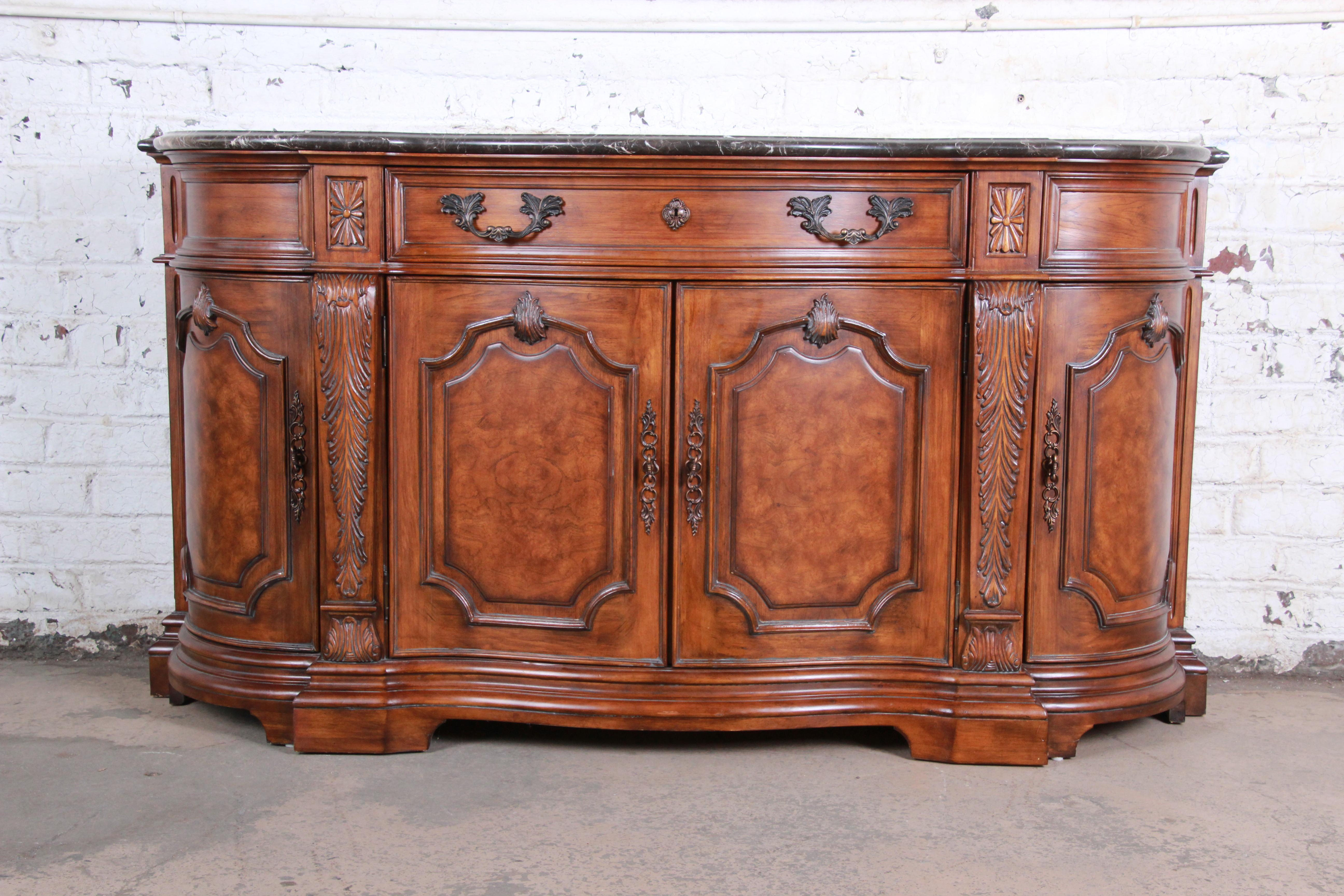 An outstanding French carved marble-top sideboard by Drexel Heritage. The sideboard features gorgeous burled walnut wood grain with nice carved wood details and a thick beveled black marble top with gray and white veining. It offers ample storage,