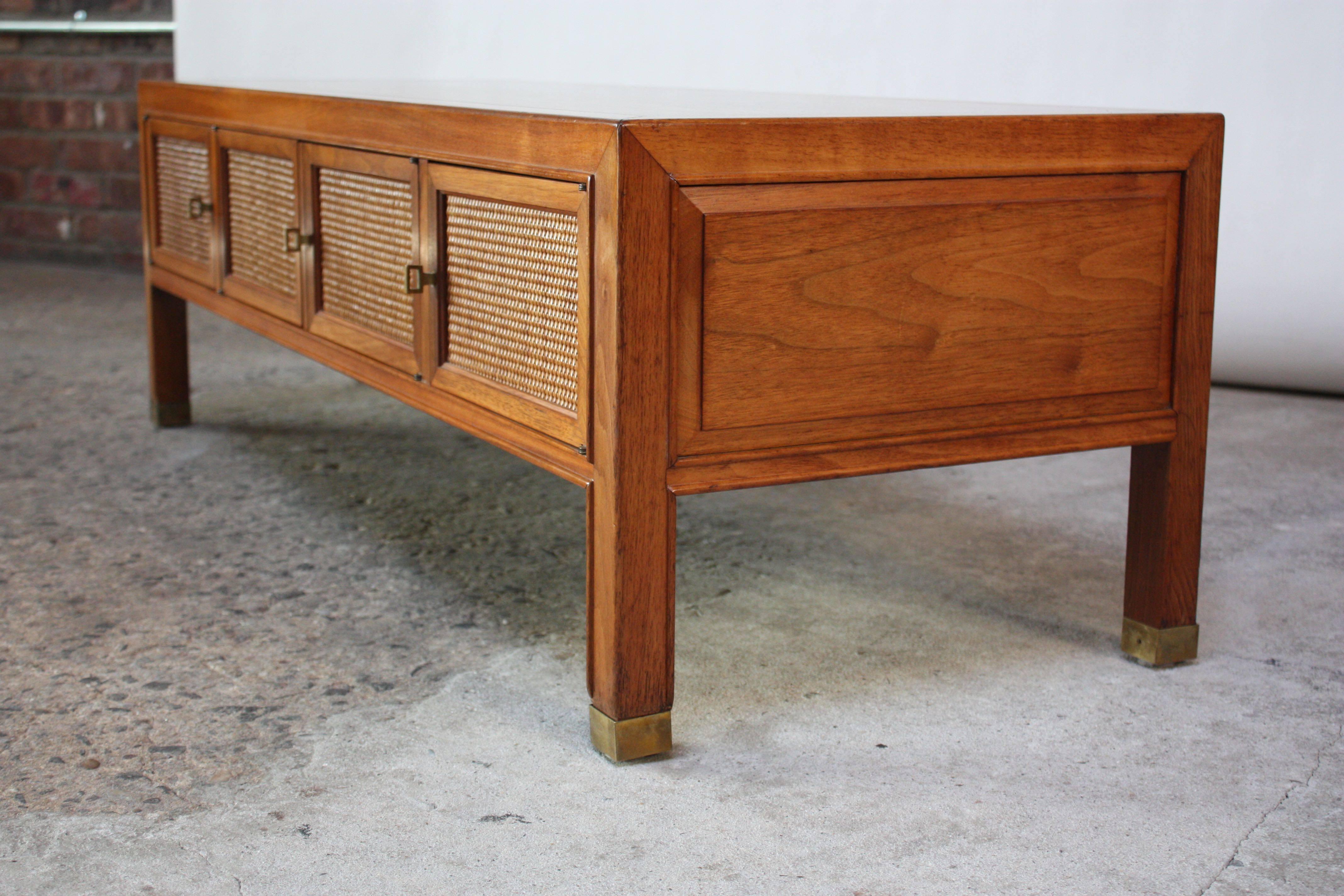 This 1960s low coffee table was designed and manufactured by Drexel for the Heritage Line.
It features four cane-front doors that all open to reveal a shared storage. Patinated brass capped feet and cabinet pulls. Finished on all sides, the back