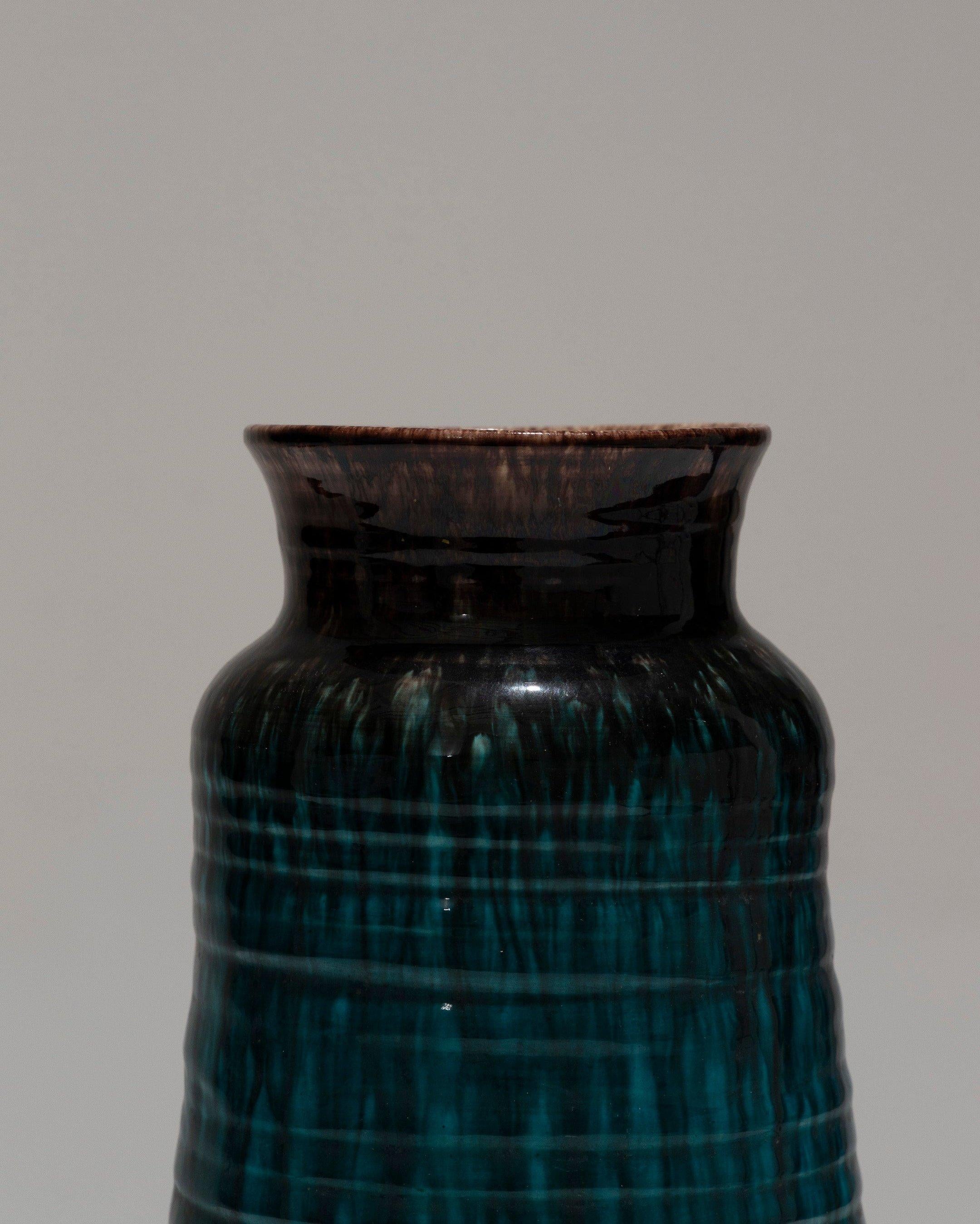 Jarrón de loza con cuerpo alto y recto y cuello acampanado, Accolay, años 50. Presenta marcas visibles de rueda y una decoración de goterones marrón hierro sobre un cuerpo turquesa oscuro. Firmado bajo la base : 'Accolay / 29.' Procedencia : Fábrica