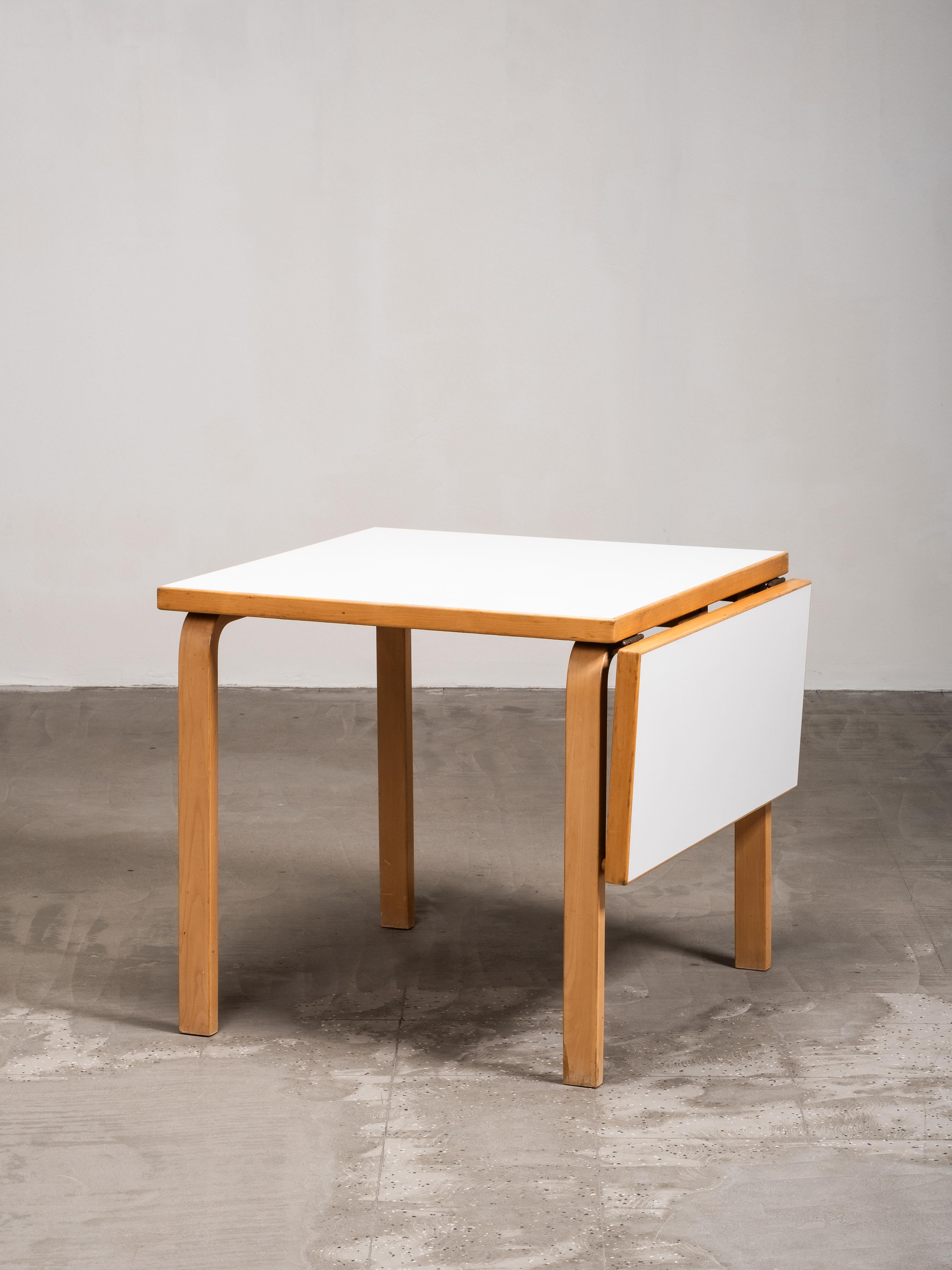 Table fonctionnelle et intemporelle à pied tombant DL82 d'Alvar Aalto, produite par Artek dans les années 1960. Le design présente les pieds en bois courbé caractéristiques d'Aalto, associés à un plateau en stratifié blanc. La construction pratique