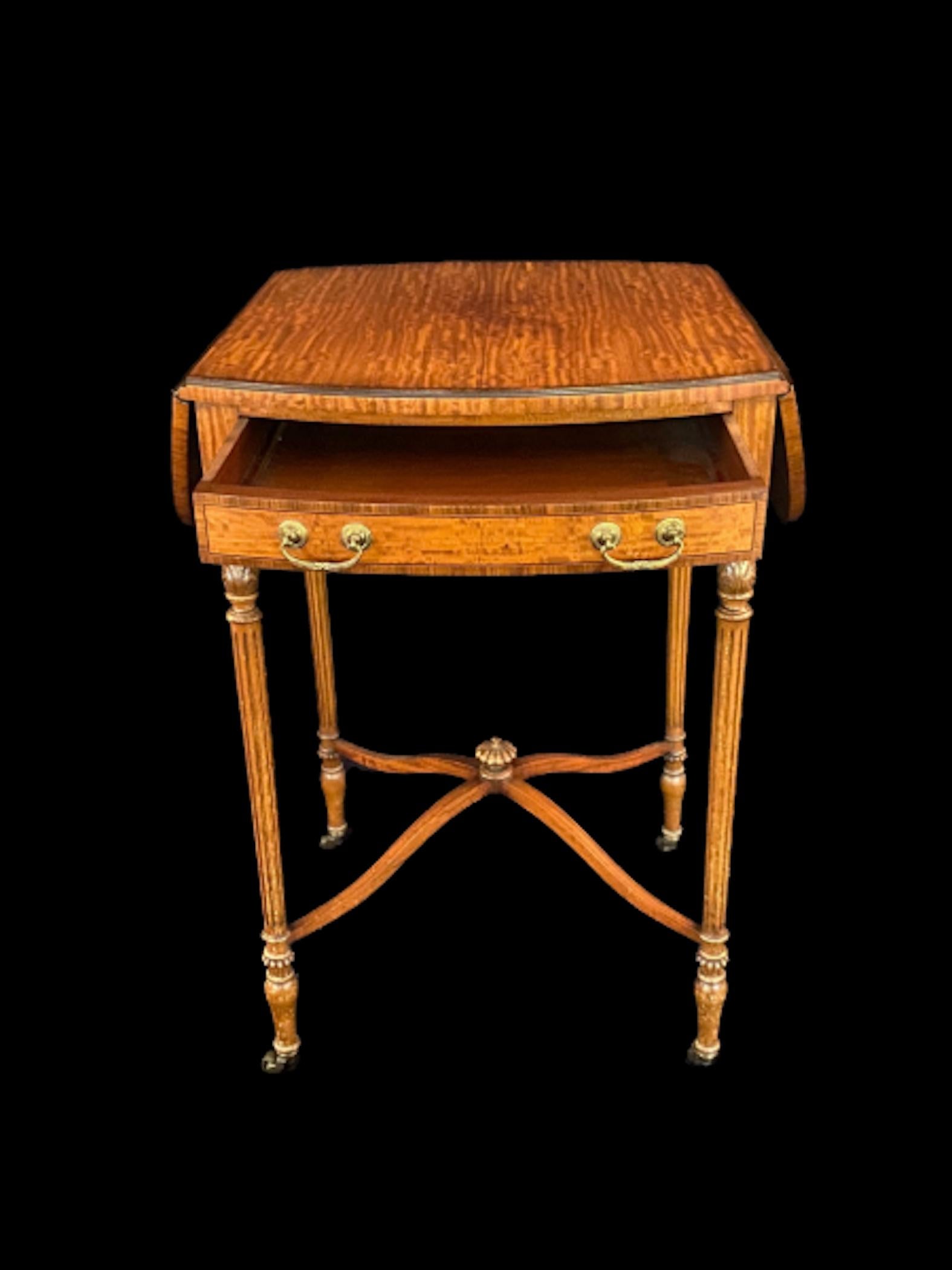 A beautiful yet delicate satinwood drop leaf oval foldable side table with remarkable natural warm tones. Standing upon four reeded legs finished with brass castors, the table also has a flush drawer is elegant brass handles. When the table top is
