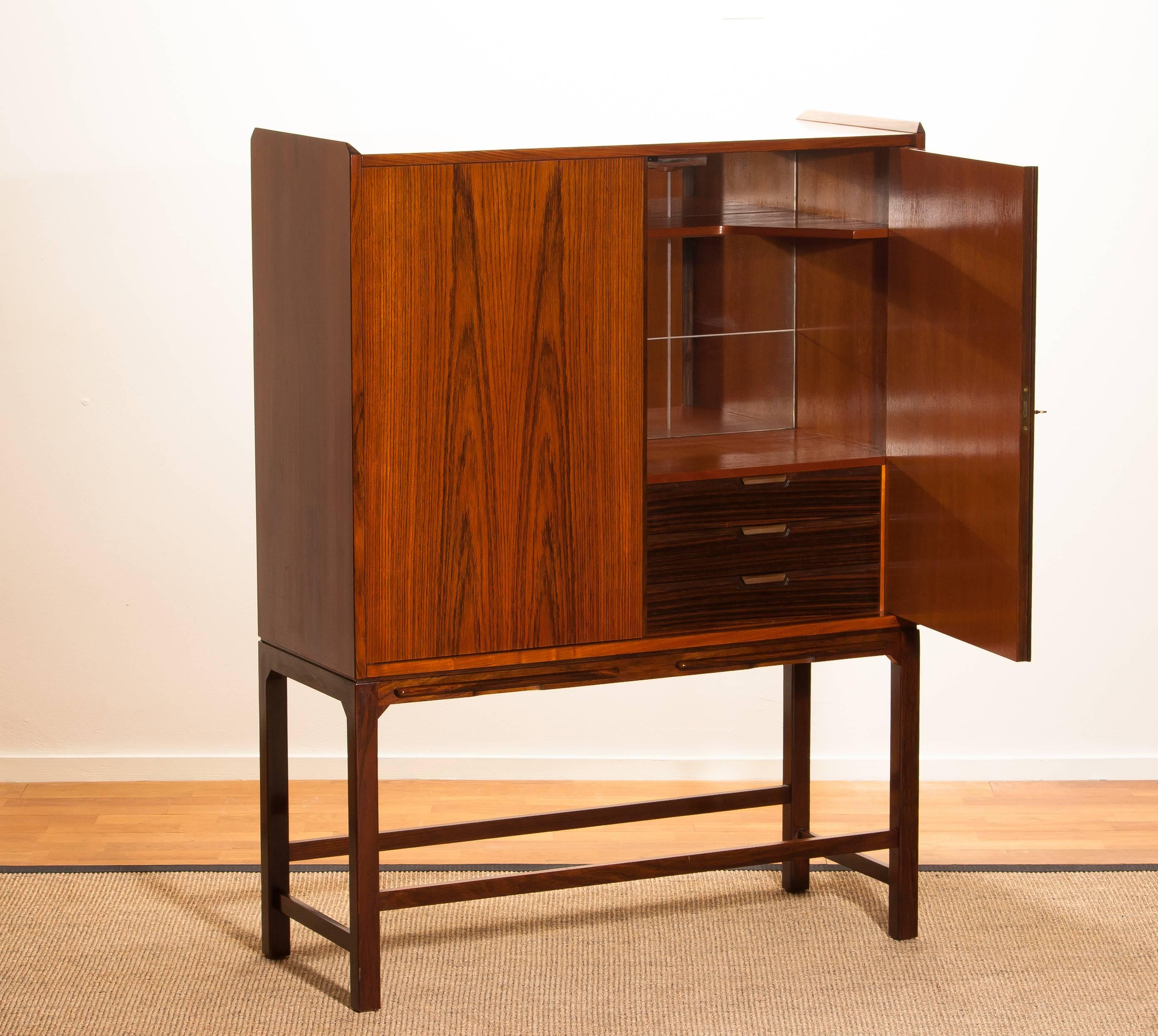 Exceptional beautiful and in perfect condition dry bar / cocktail cabinet of mahogany on a skinny rosewood stand. In the rosewood stand are (also in rosewood) two extendable shelfs.
Inside the cabinet is made of teak. The three drawers are in