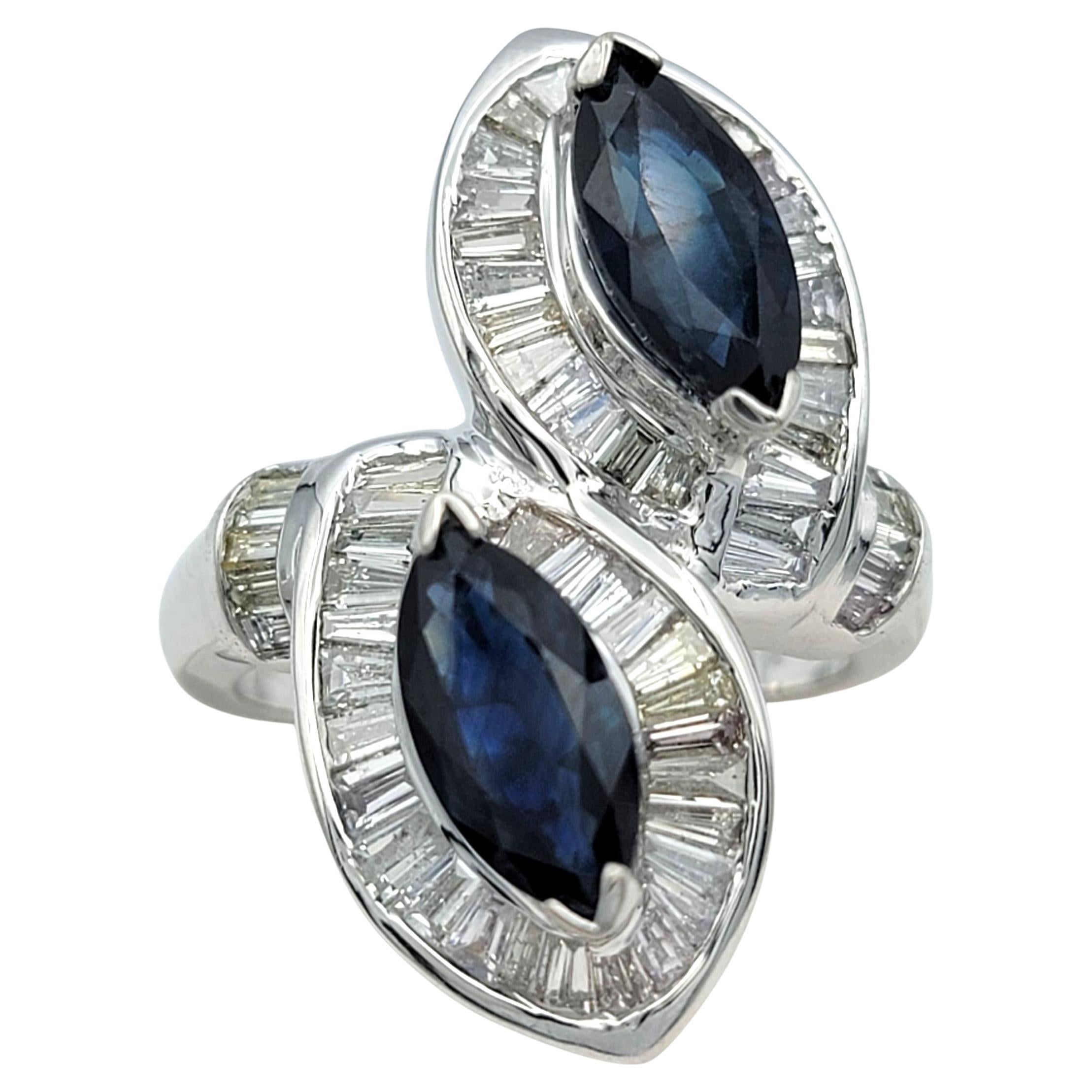 Dieser auffällige Saphir- und Diamantring zeigt zwei tiefblaue Marquise-Saphire, die sich in einem dramatischen Bypass-Design gegenüberstehen. Jeder Saphir wird von einem Halo aus Baguette-Diamanten umrahmt, die in ihrer Brillanz schimmern und den