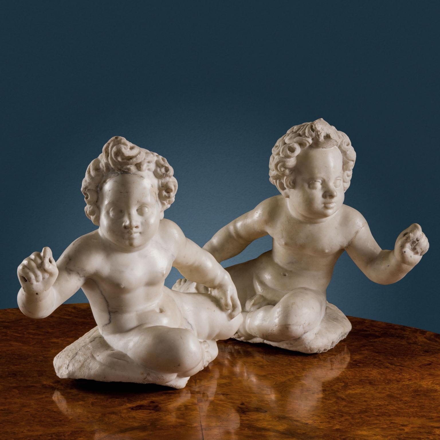 Ein Putti-Paar aus Carrara-Marmor, dargestellt in spiegelbildlicher Haltung: locker liegend, die Büste vorgerückt und leicht seitlich abgewinkelt, ein Arm auf dem Oberschenkel ruhend, während der andere Arm erhoben ist und die geschlossene Hand ein