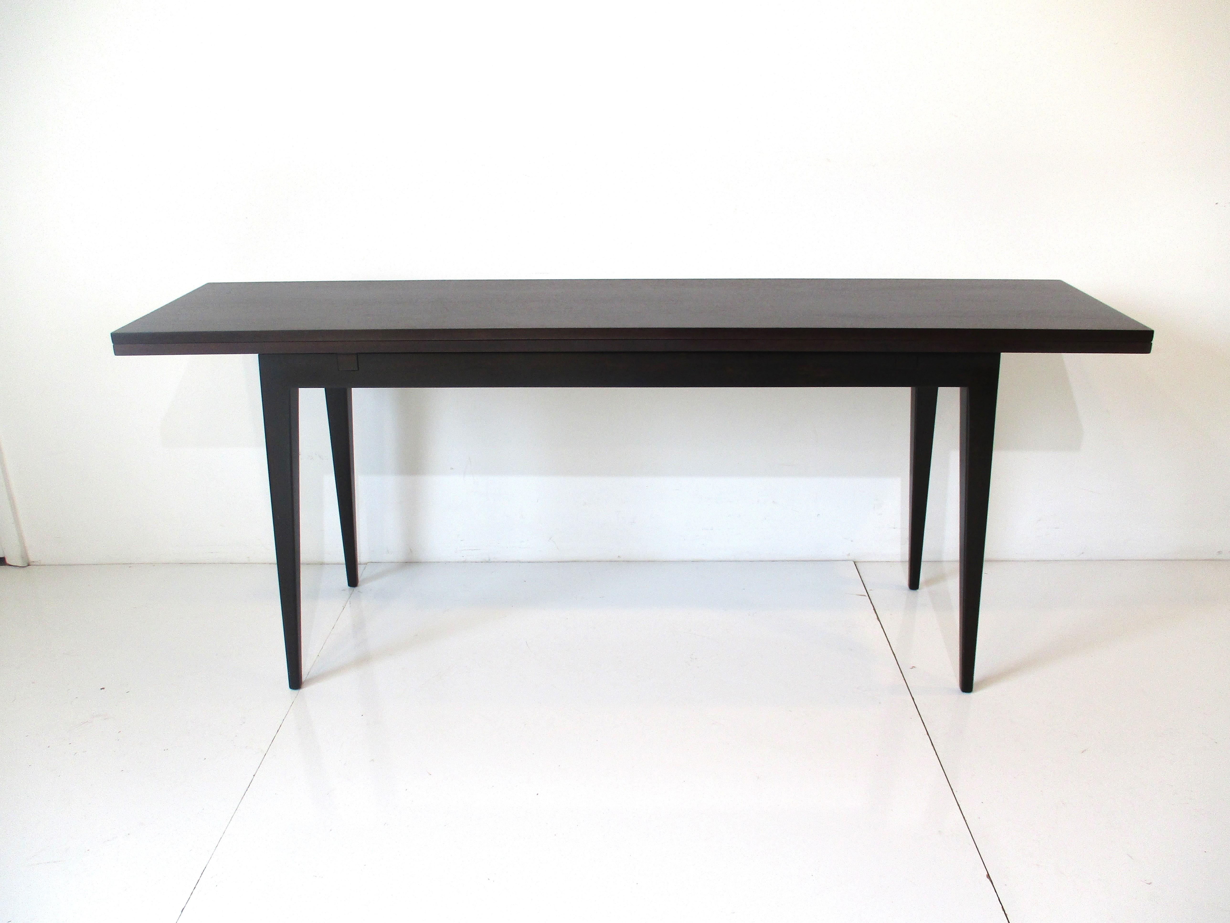 A well crafted and rich looking console flip top dining table with dark ebony finish and satin black tapered legs. The back edge of the piece has hinges and when the top is pulled forward and the upper panel is opened it makes a nice sized dining
