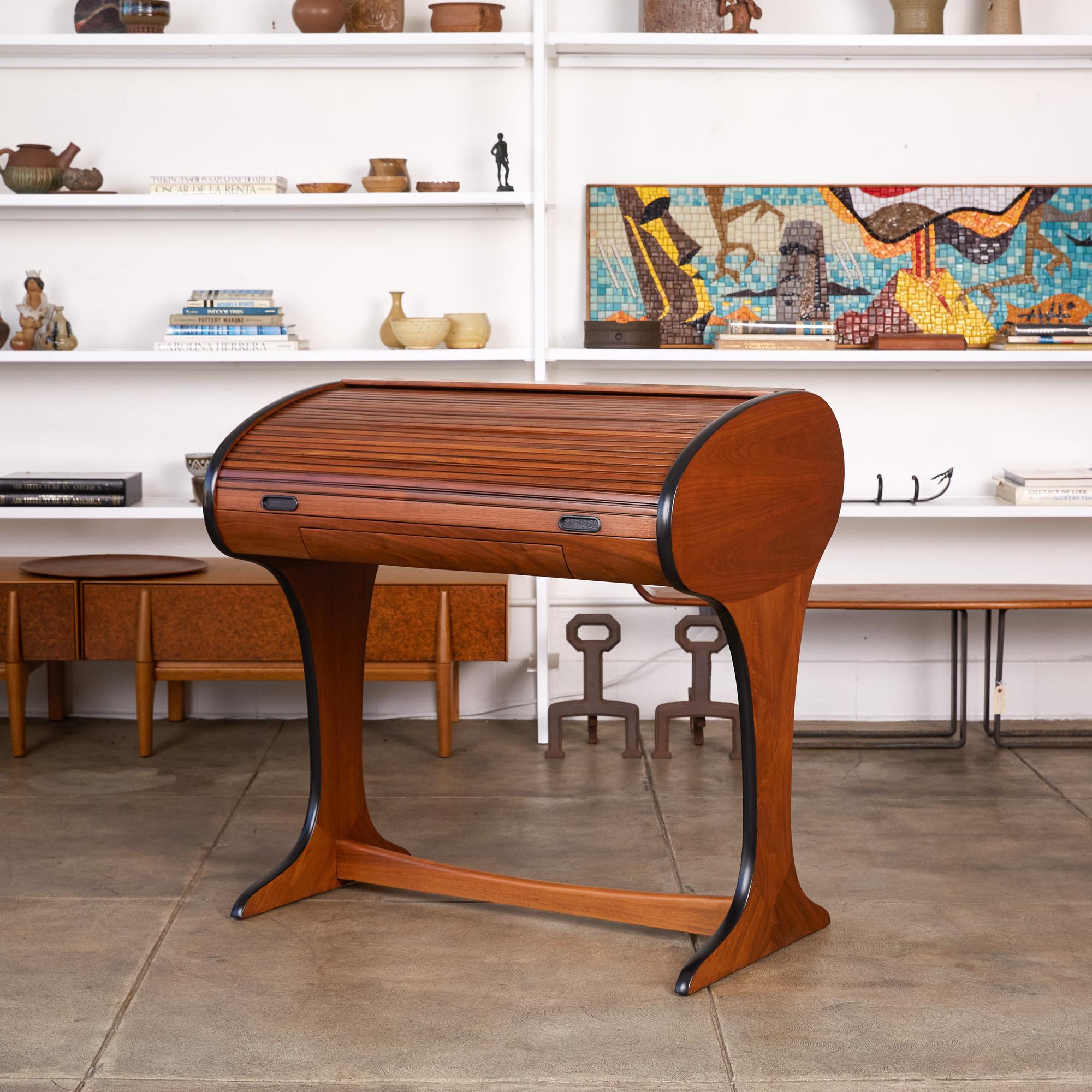 Dunbar style roll top writing desk. The desk features a walnut frame and tambour door, with black painted trim. The slatted wood pattern of the tambour door is repeated on the reverse side of the body. The sculpted walnut legs flow seamlessly from