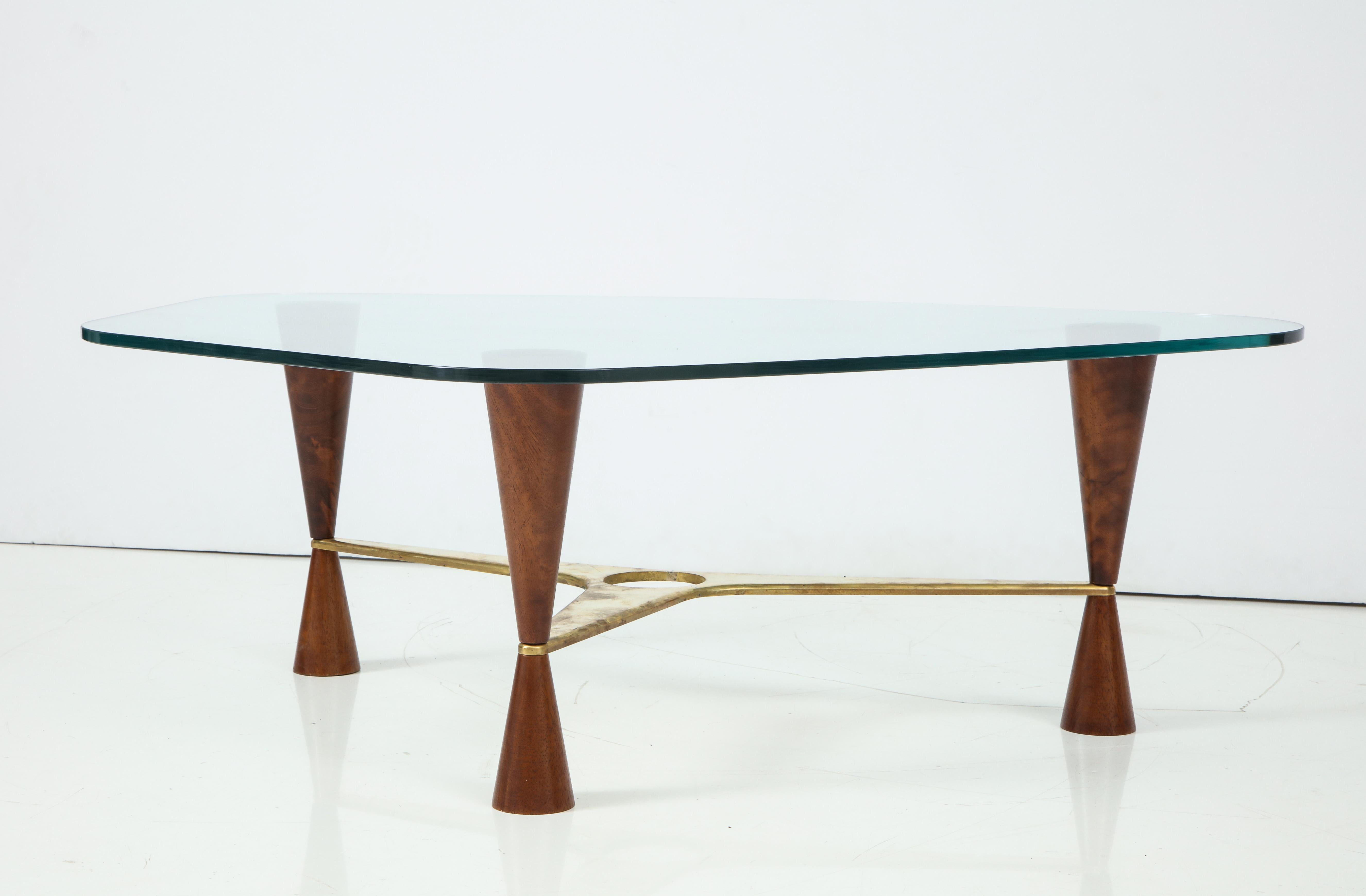 A Dunbar style cocktail or coffee table with three walnut conical legs tapered two-thirds down where the pierced shaped brass stretcher is anchored, surmounted by a polygonal shaped glass top with rounded edges.