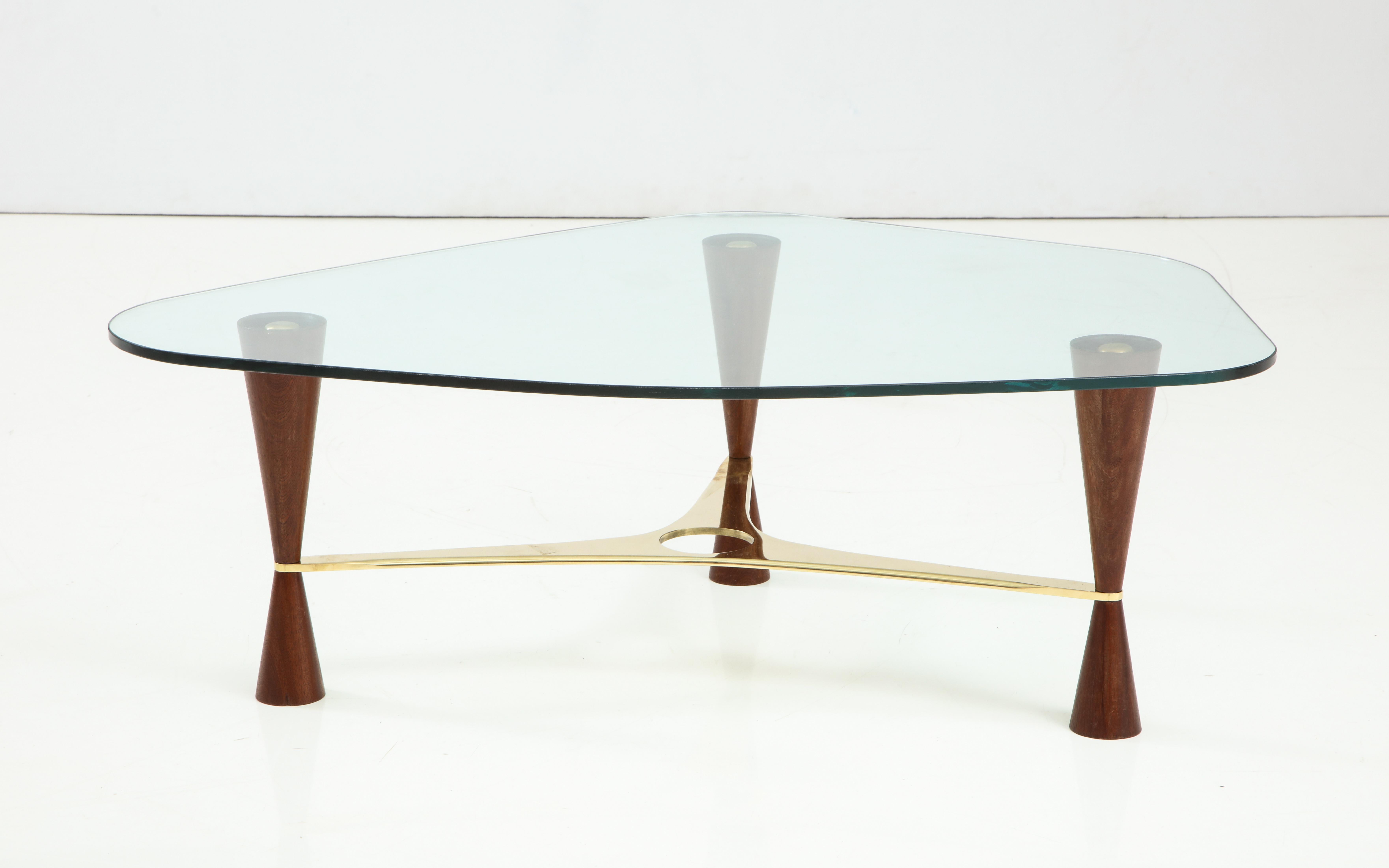 Italian contemporary cocktail table with three walnut conical legs tapered two-thirds down where the pierced shaped brass stretcher is anchored, surmounted by a polygonal shaped glass top with rounded edges.