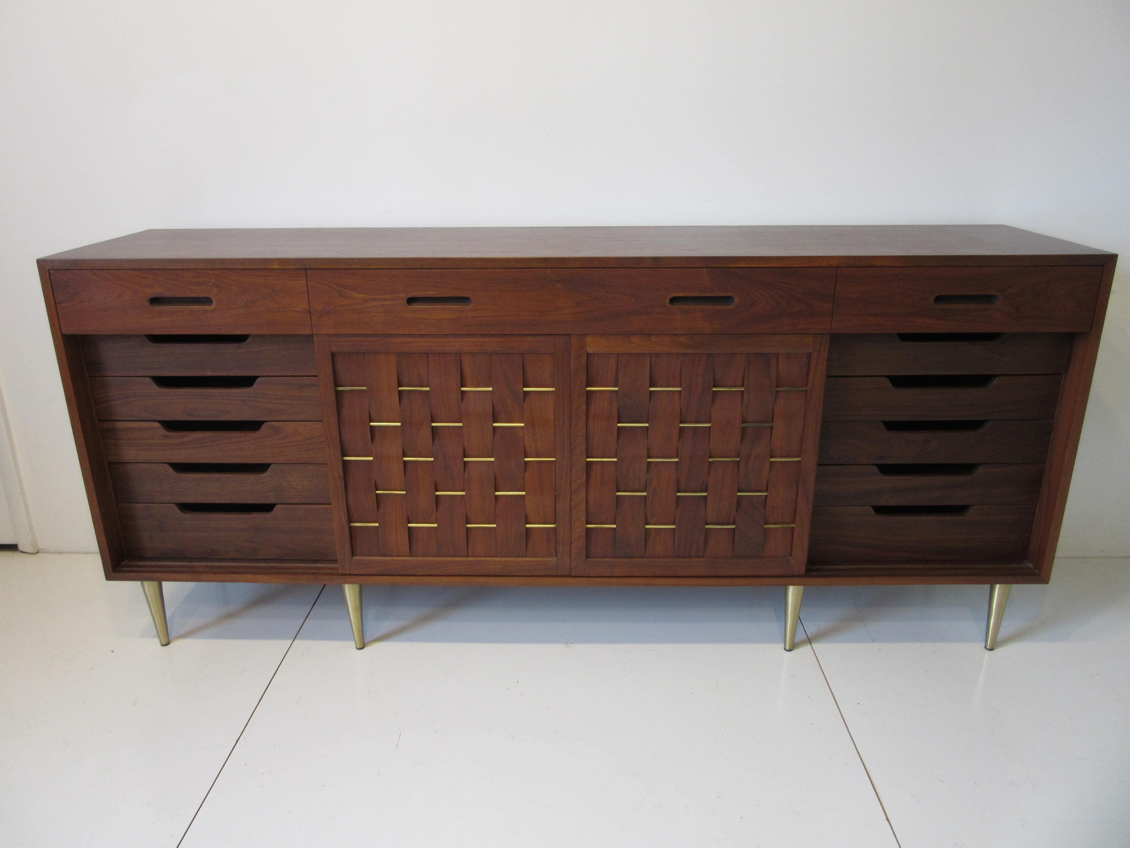 A beautiful medium toned walnut credenza sideboard with four sliding doors having interwoven strips of walnut with brass details. The three top drawers have built in pulls and behind the sliding doors each end has additional drawers with removeable