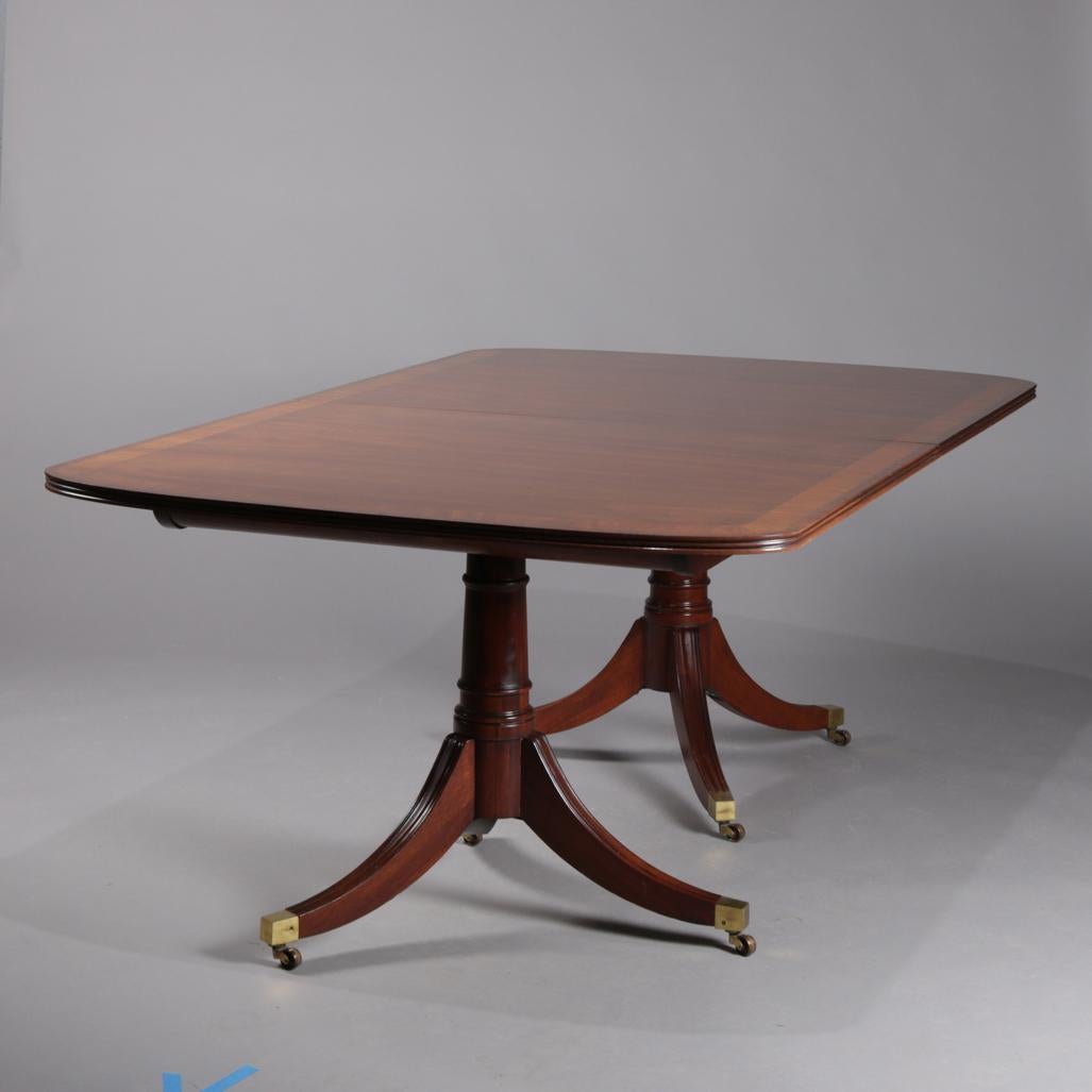 Duncan Phyfe School dining table by White Furniture Co. features mahogany construction with top having crossbanding, raised on double tripod pedestals with reeded legs terminating in bronze caps on casters, including three matching leaves, circa