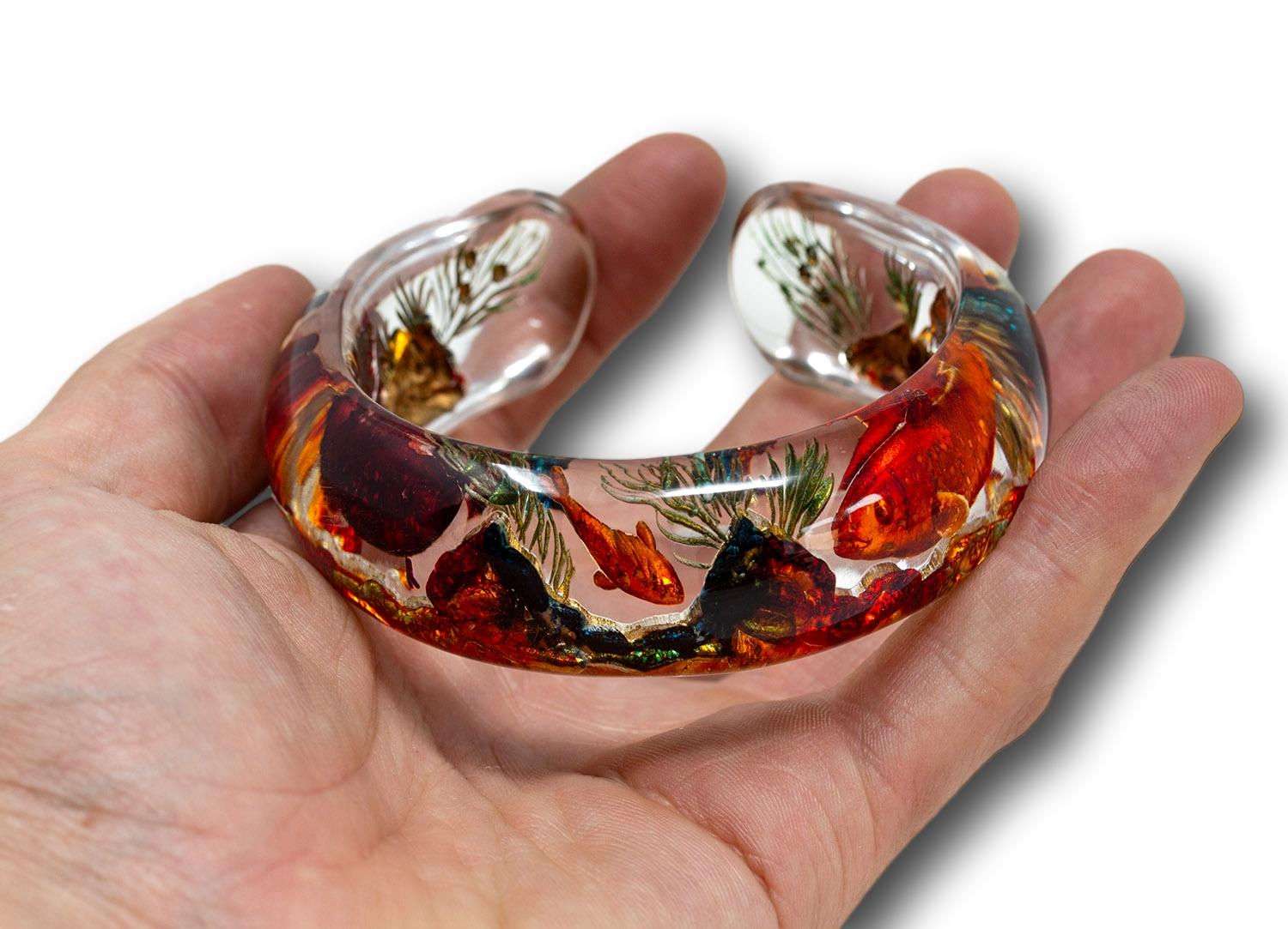 Matching the Dunhill Aquarium Lighter (His & Hers) 

The bangle formed in lucite features a reverse decorated central sea scape scene with fish amongst foliage and rock work in beautiful orange and golden colours giving it a striking vibrant