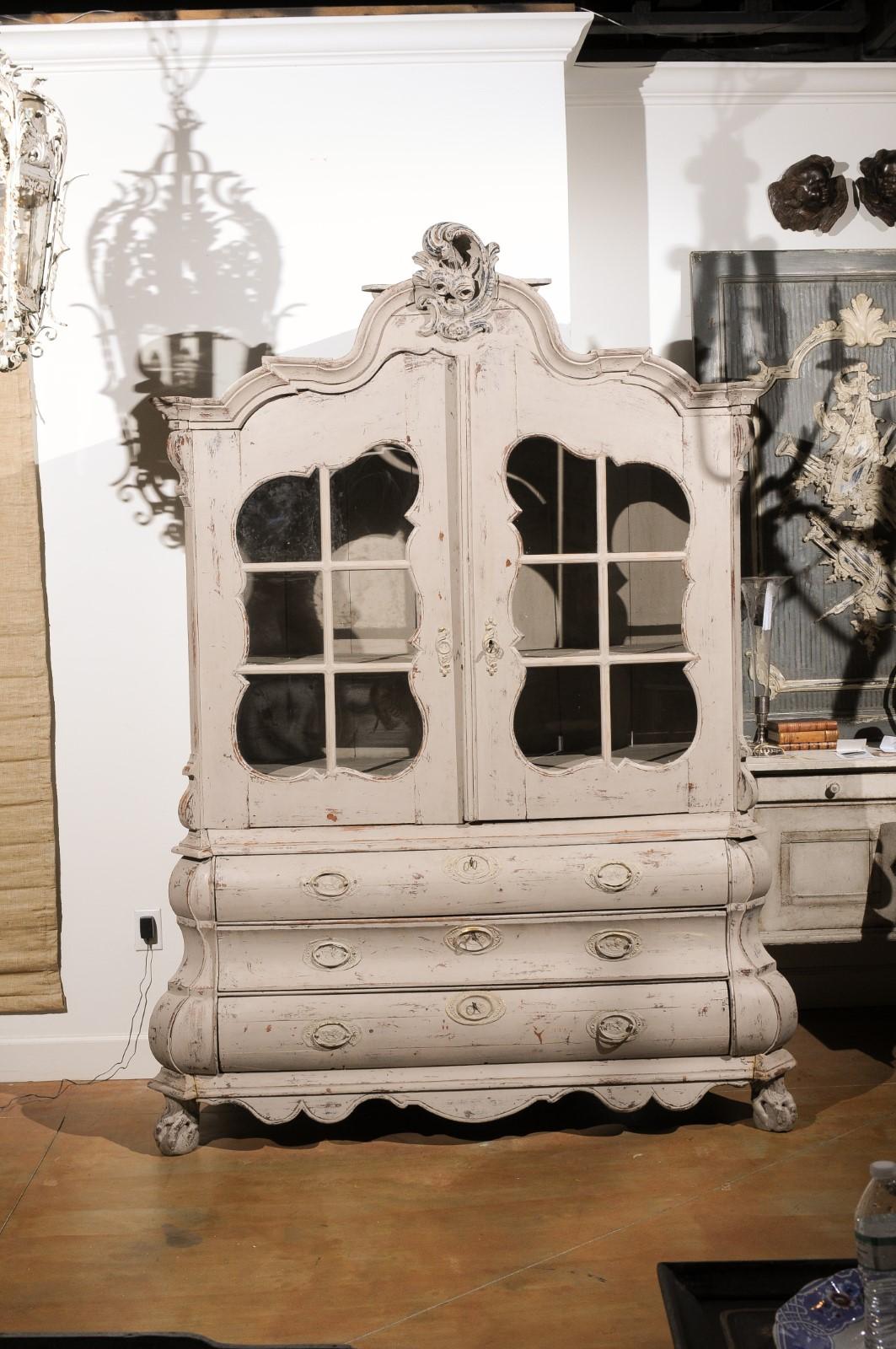 A Dutch Rococo Revival painted cabinet from the mid 19th century with two glass doors over three drawers and original hardware. This painted cabinet, created in the Netherlands during the 1850s, features a bonnet shaped pediment, exquisitely adorned
