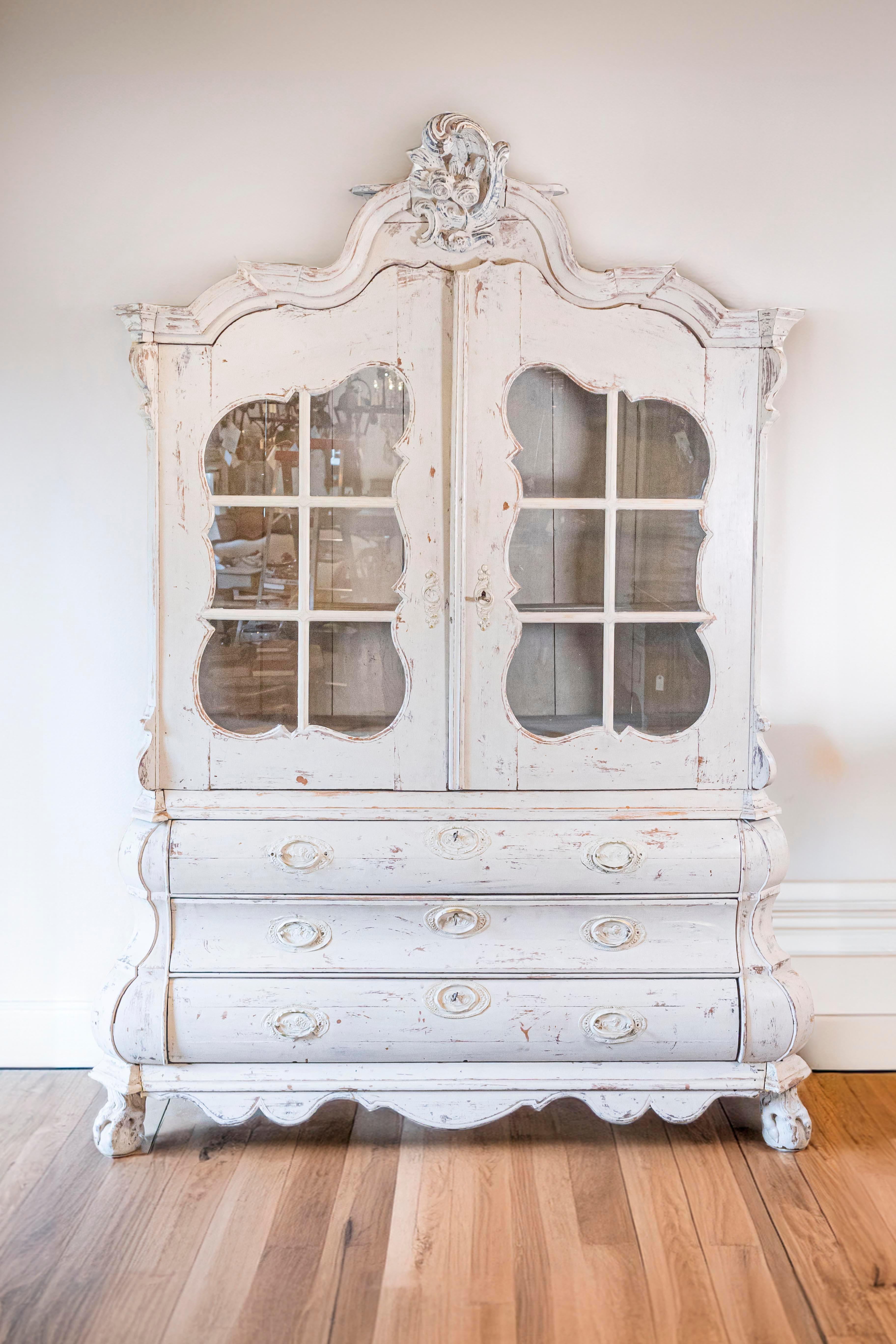 A Dutch Rococo Revival painted cabinet from the mid 19th century with two glass doors over three drawers and original hardware. This painted cabinet, created in the Netherlands during the 1850s, features a bonnet shaped pediment, exquisitely adorned