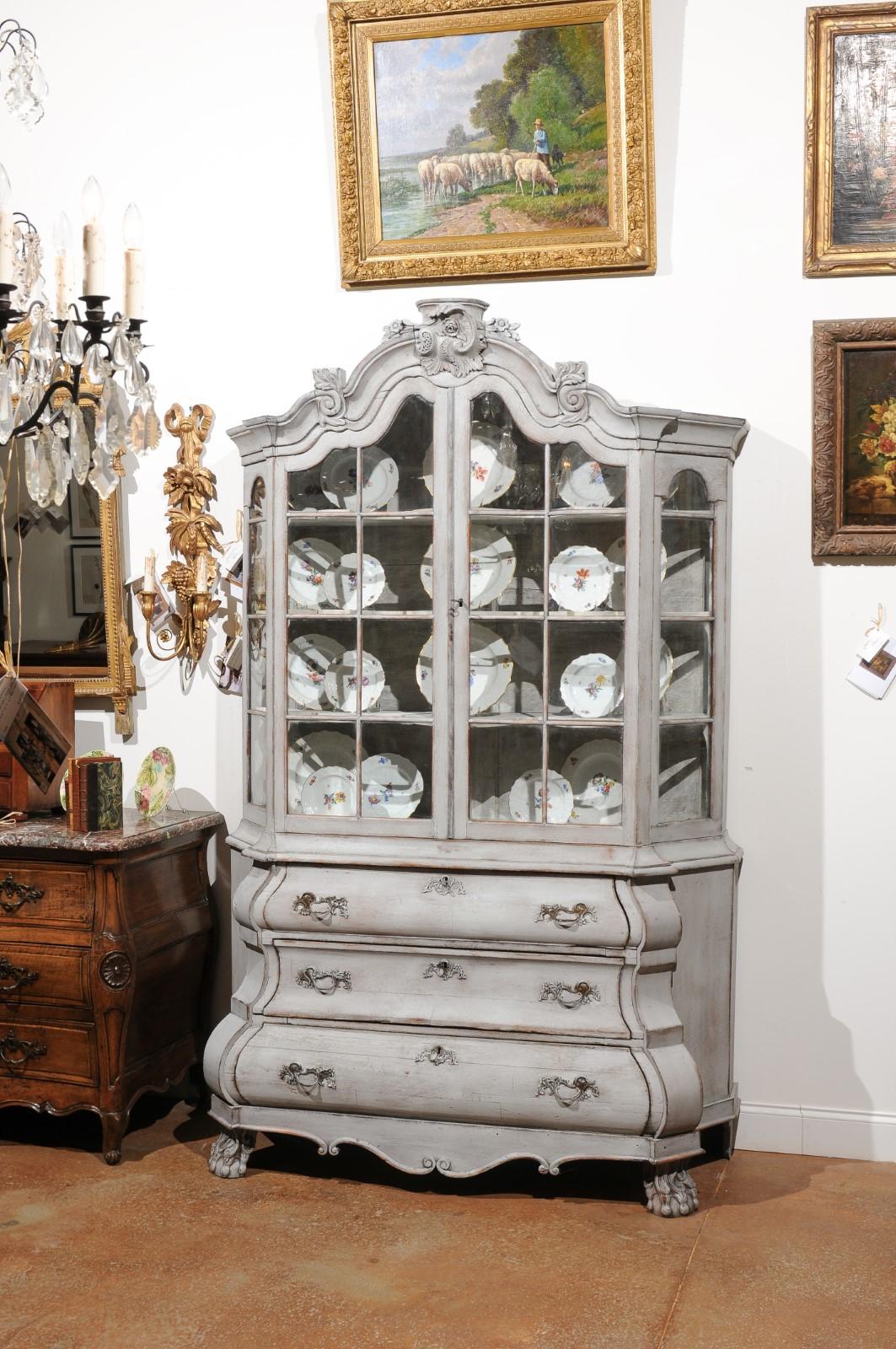 A Dutch Rococo style painted wood vitrine cabinet from the 19th century, with glass doors, rocaille-carved crest and bombé chest. Born in the Netherlands during the 19th century, this painted vitrine features an eye-catching molded cornice, adorned