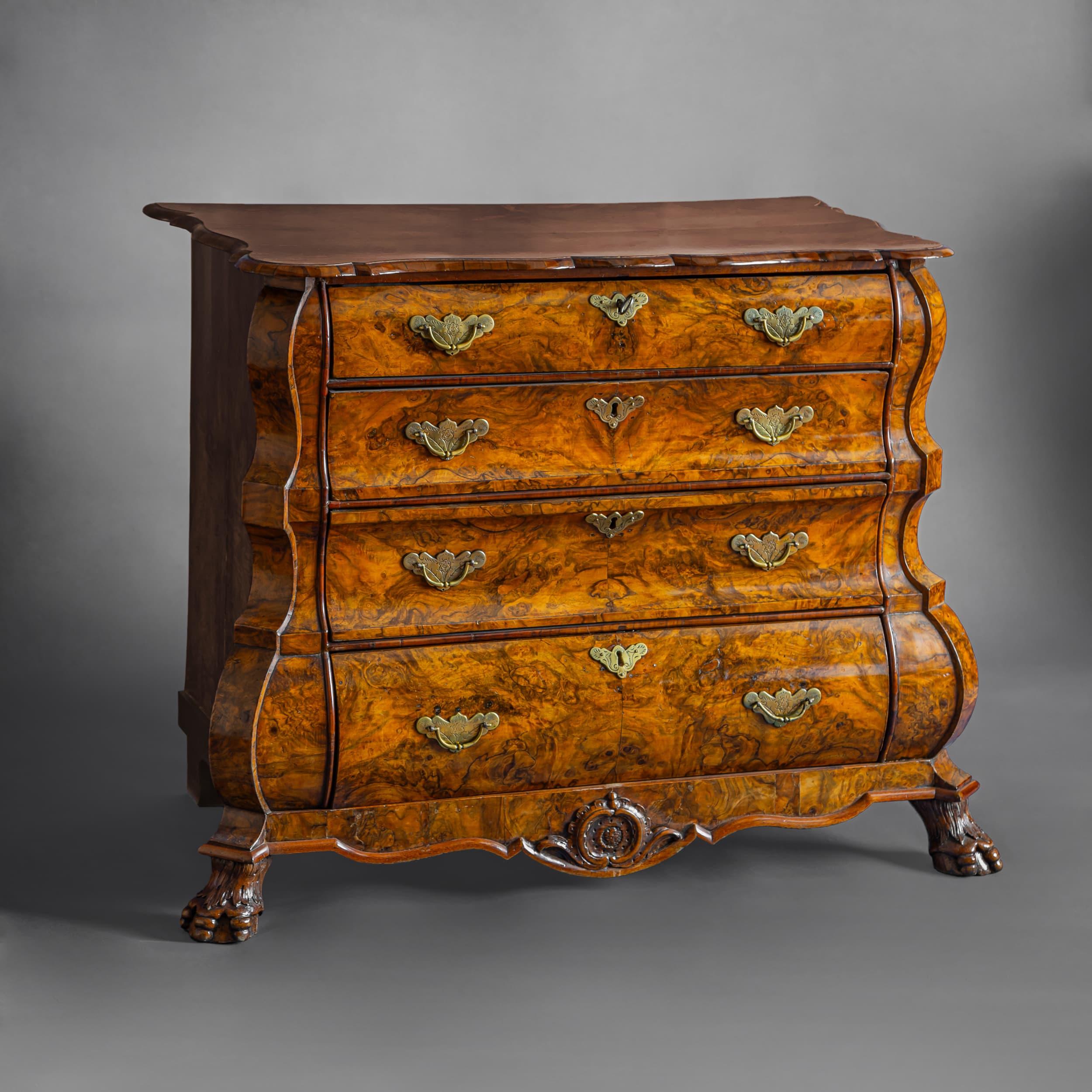 Dutch four-drawer Baroque-style chest in trapezoidal form, featuring the characteristic serpentine front and sides, carved claw feet, and rocaille decorations on the curved apron. The drawer fronts are adorned with chiseled, openwork brass fittings.