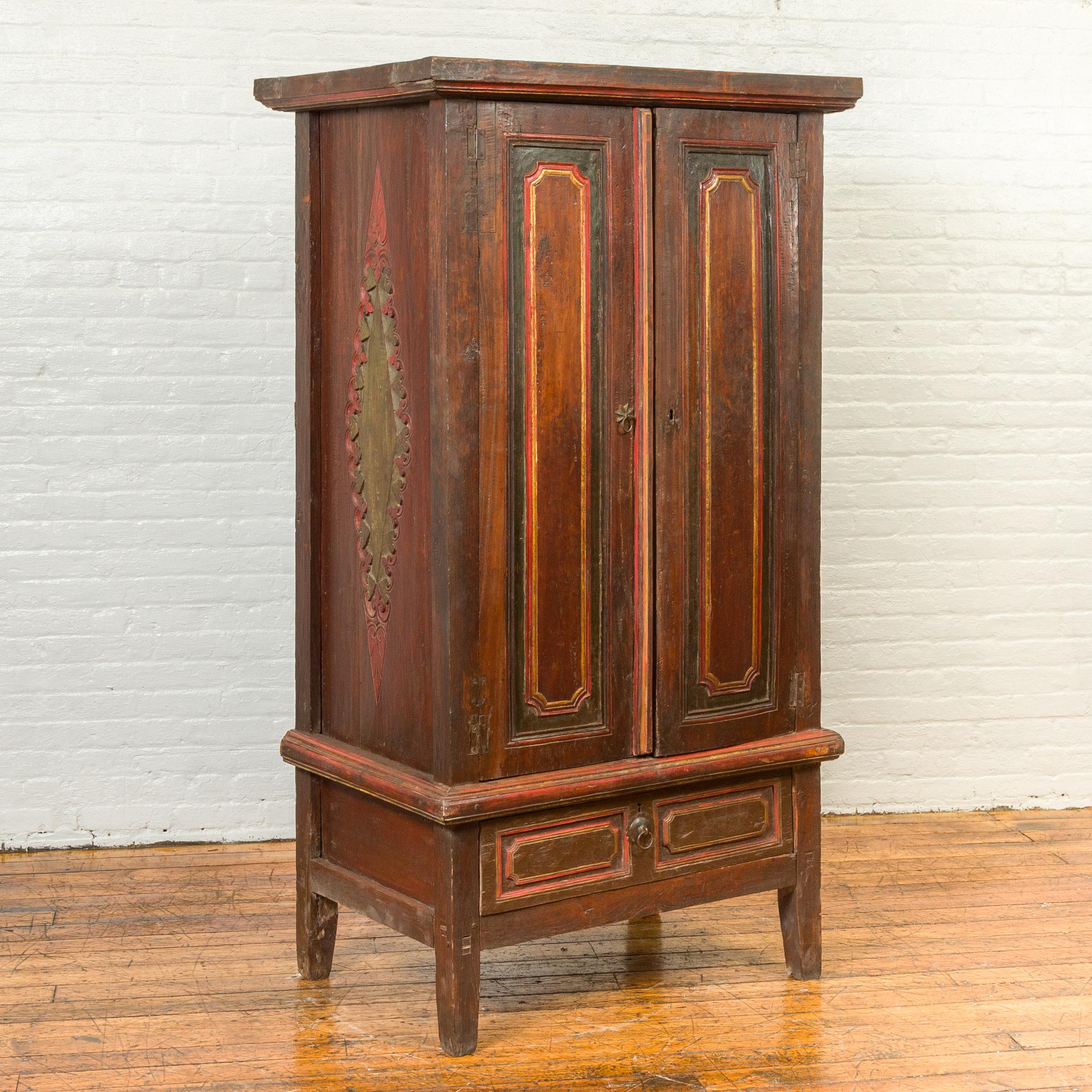 A Dutch colonial wooden cabinet from the 19th century, with polychrome finish, single drawer and carved sides. Created in Indonesia during the 19th century, this tall cabinet features a cornice overhanging two large doors, each adorned with a green,