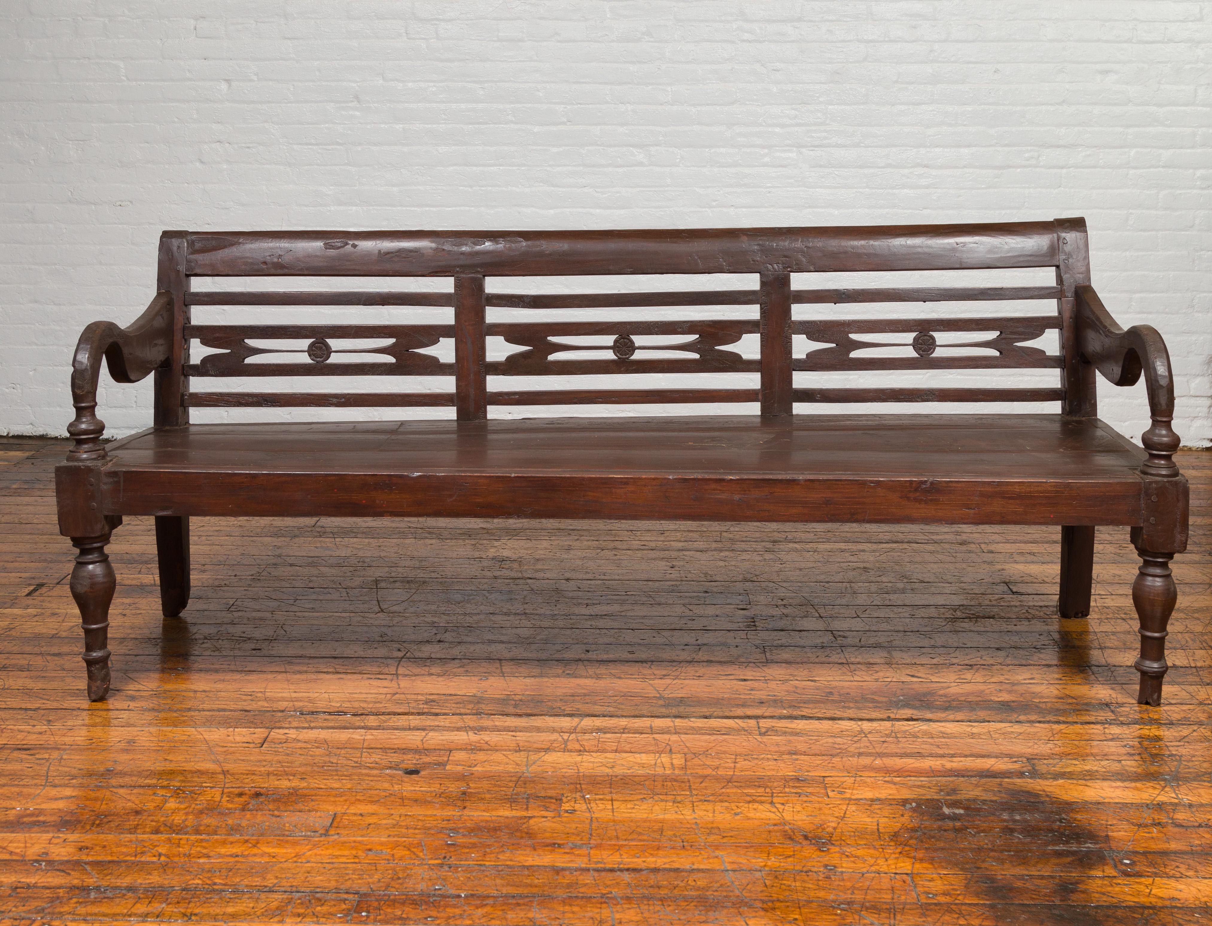 An antique Dutch Colonial Indonesian teak wood garden bench from the 19th century, with pierced back, petite medallions and scrolling arms. Found in Jakarta, this 19th century Dutch Colonial bench features a slightly out-curving back, pierced with