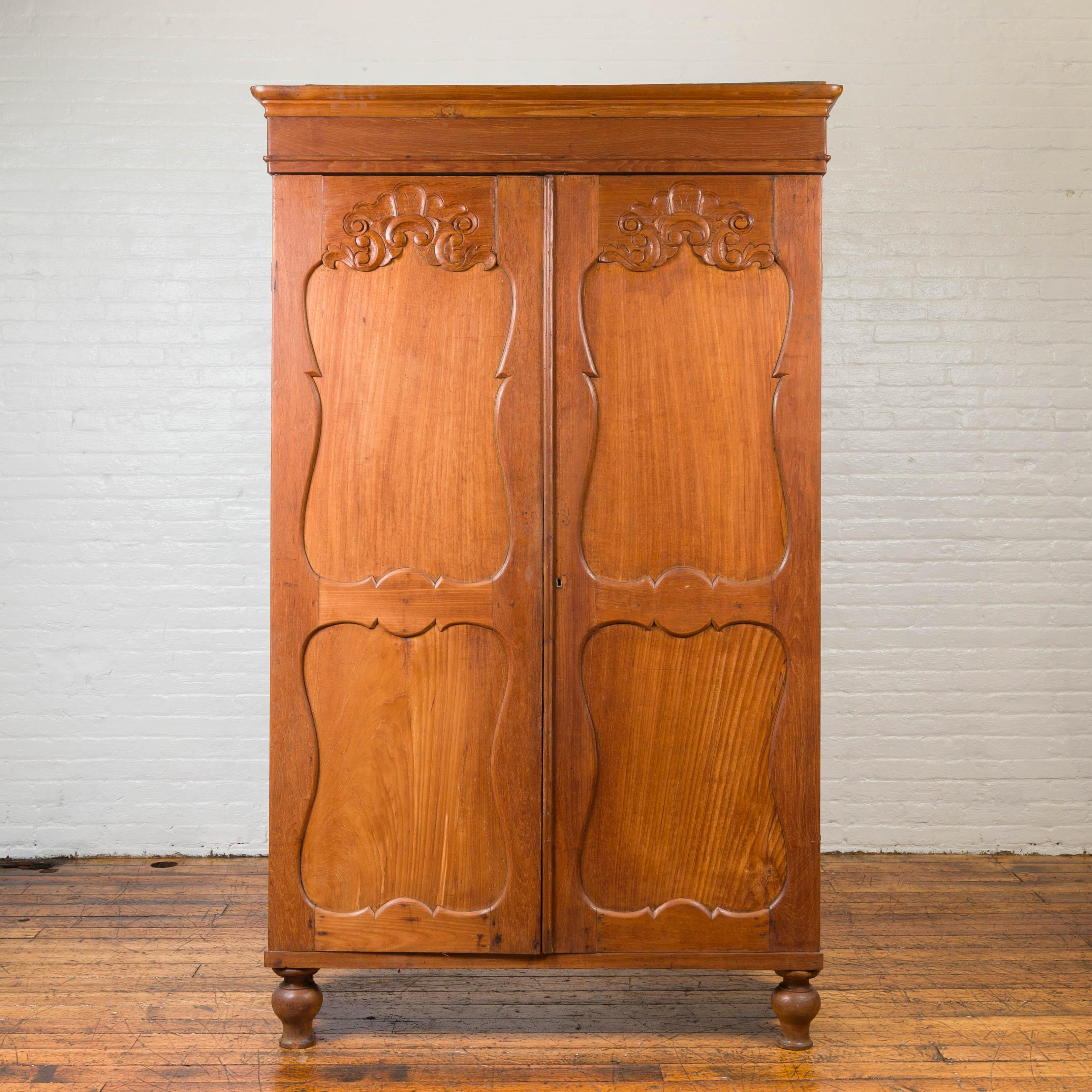 A 19th century Dutch Colonial teak cabinet with carved panels and inner shelves. Born in Indonesia during the 19th century, this tall teak cabinet features a beveled cornice sitting above two large doors, adorned with recessed panels topped with