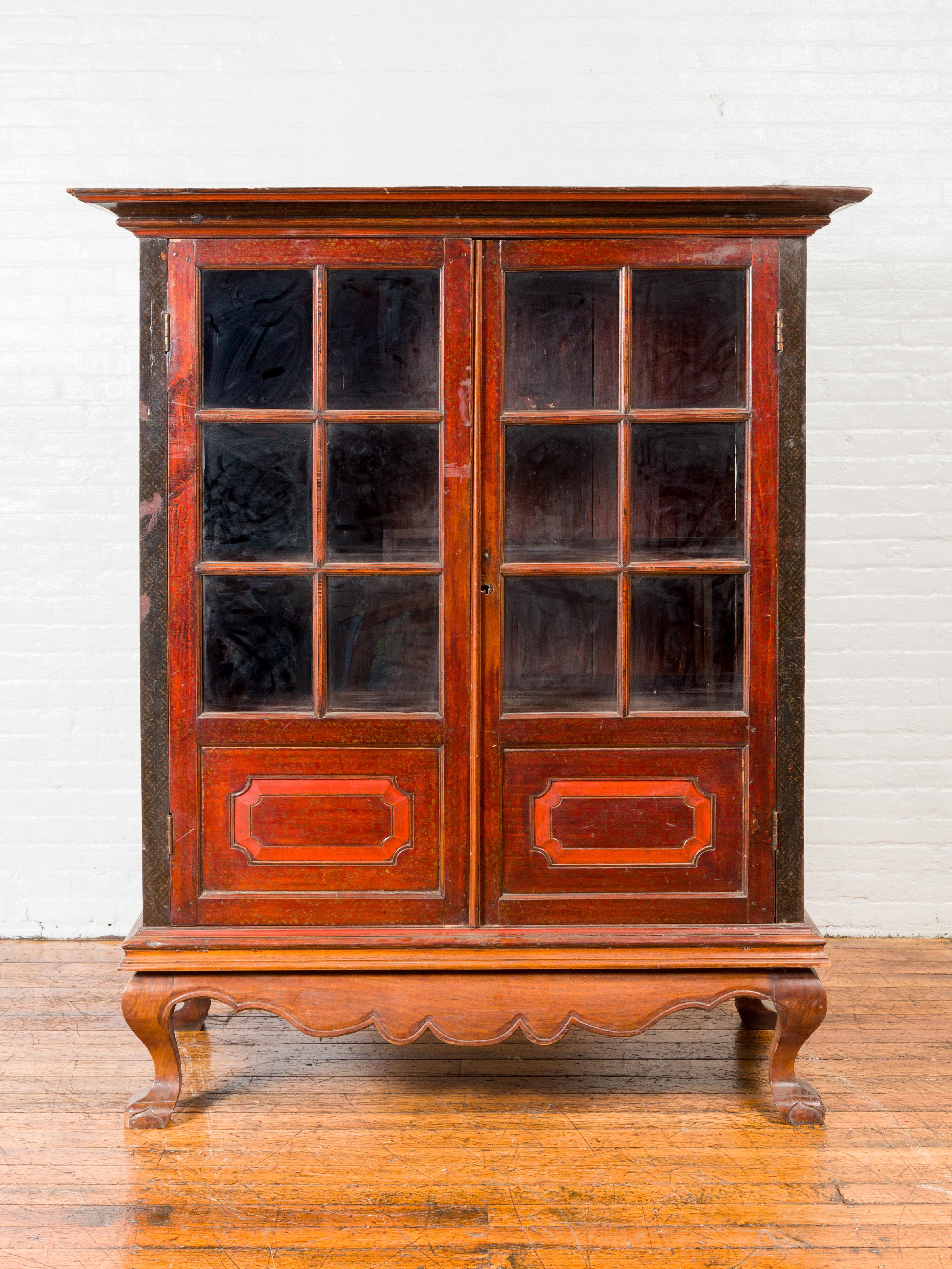 A Dutch Colonial Indonesian antique lacquered wooden cabinet from the early 20th century, with paneled glass doors, carved apron and claw feet. Crafted during the Dutch Colonial period, this tall cabinet features a molded cornice overhanging two