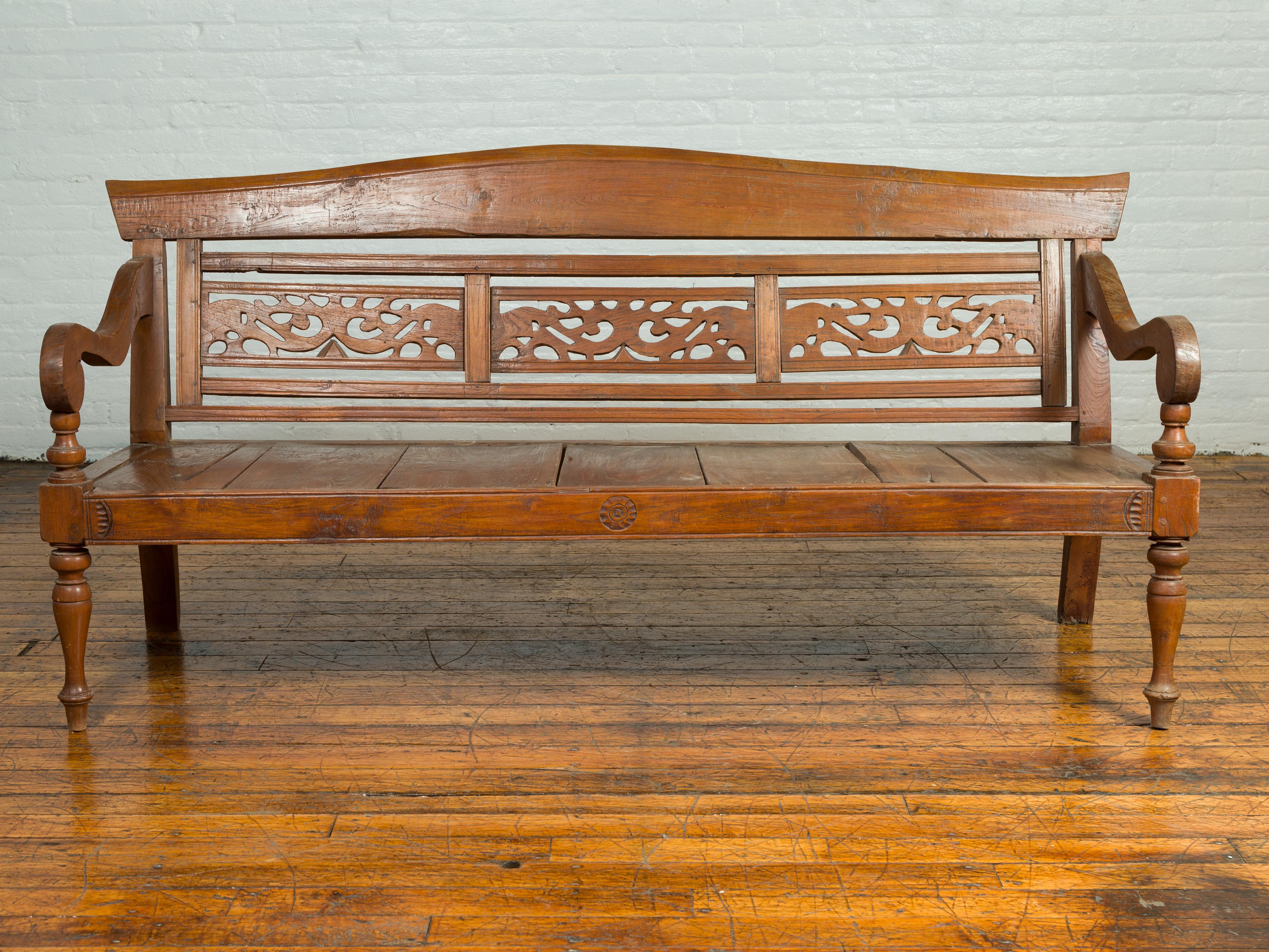 An antique Dutch Colonial Indonesian wooden bench from the early 20th century, with carved back and voluted arms. Born in Indonesia during the early years of the 20th century, this Dutch Colonial bench features a slightly slanted camelback, adorned