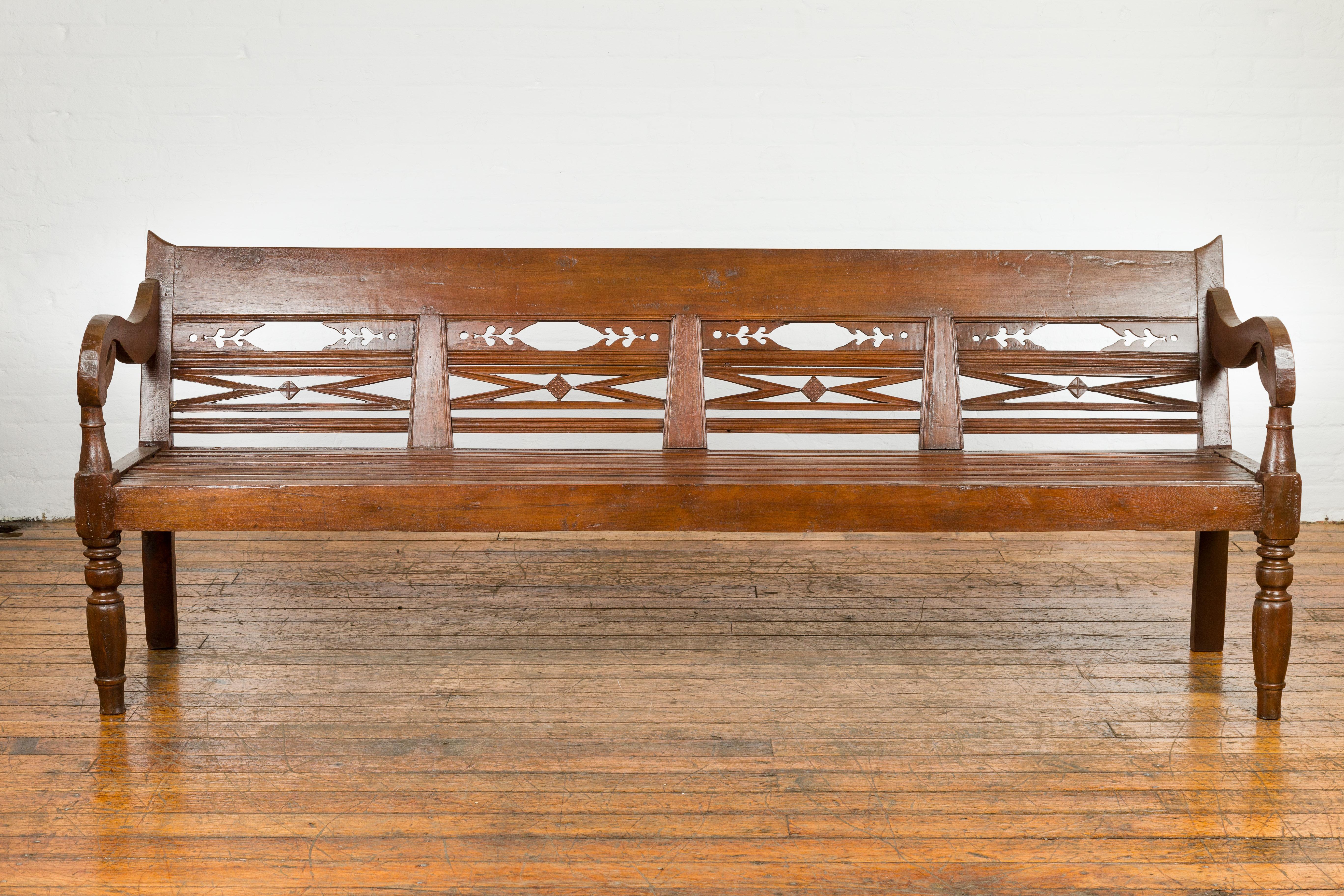 A Dutch Colonial wooden bench from the early 20th century, with carved back, scrolling arms and turned baluster legs. This early 20th-century Dutch Colonial wooden bench is a testament to exquisite craftsmanship and timeless design, gracefully