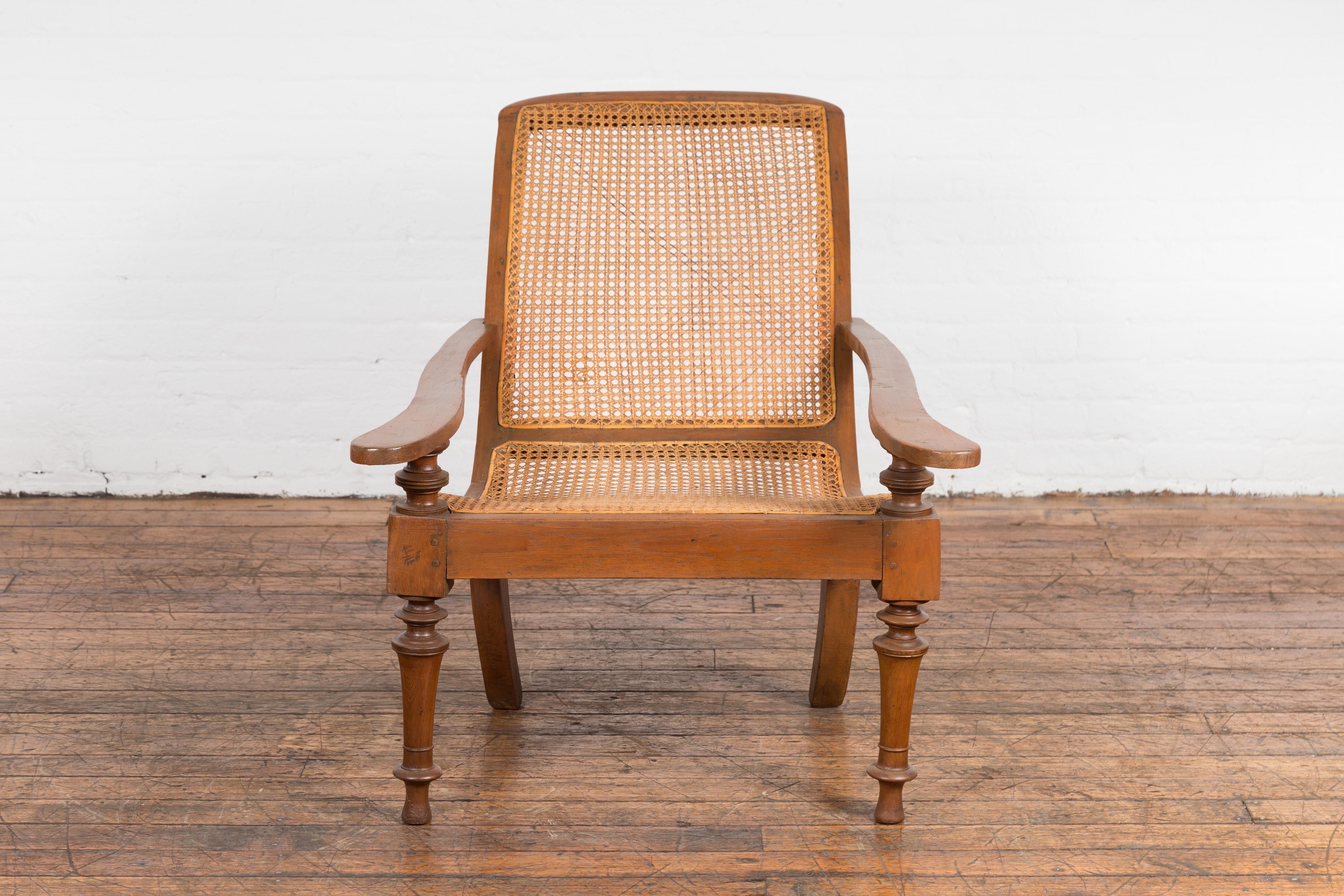 An antique Dutch Colonial Indonesian cane and wood plantation chair from the 20th century with extending arms and turned baluster legs. Enhance your living space with the charm and exotic allure of this antique Dutch Colonial Indonesian plantation