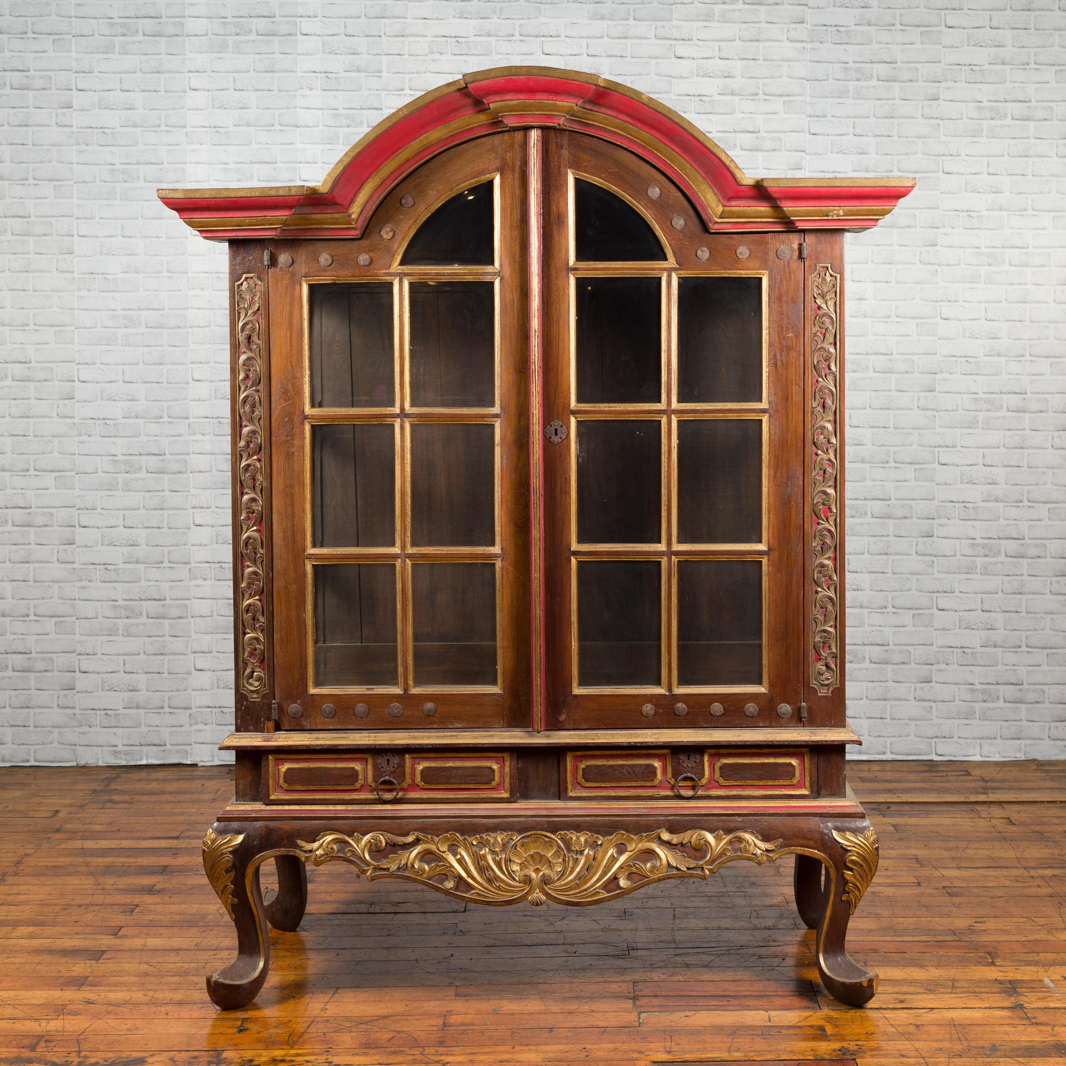 A Dutch Colonial cabinet with bonnet top, glass doors and red and gilded accents. This early 20th-century Dutch Colonial cabinet is a stunning representation of period craftsmanship, featuring a graceful 