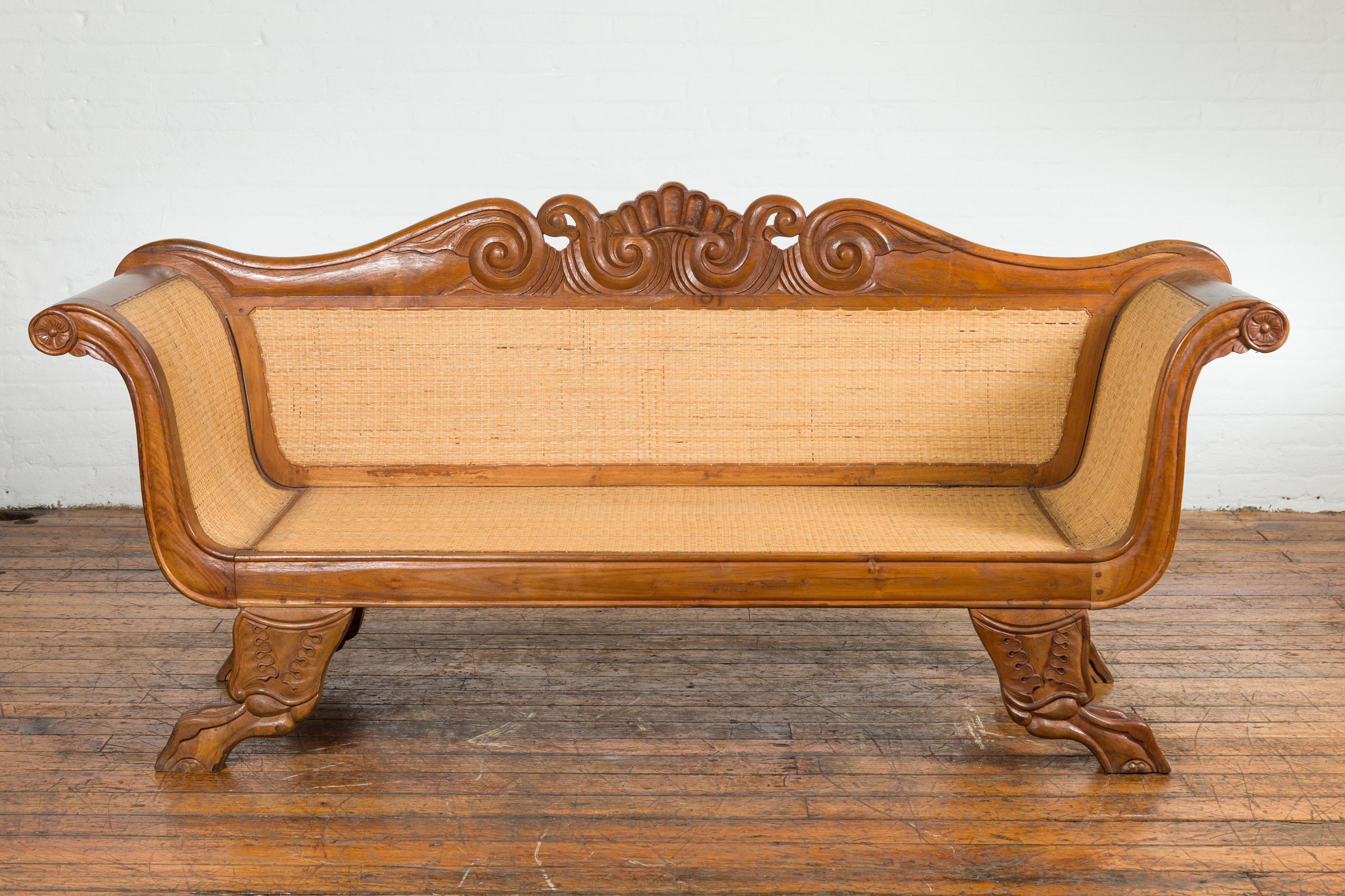 An antique Dutch Colonial period Javanese teak wood settee from the early 20th century, with inset woven rattan, carved back, large out-scrolling arms and curving legs. Created on the Island of Java during the early years of the 20th century, this