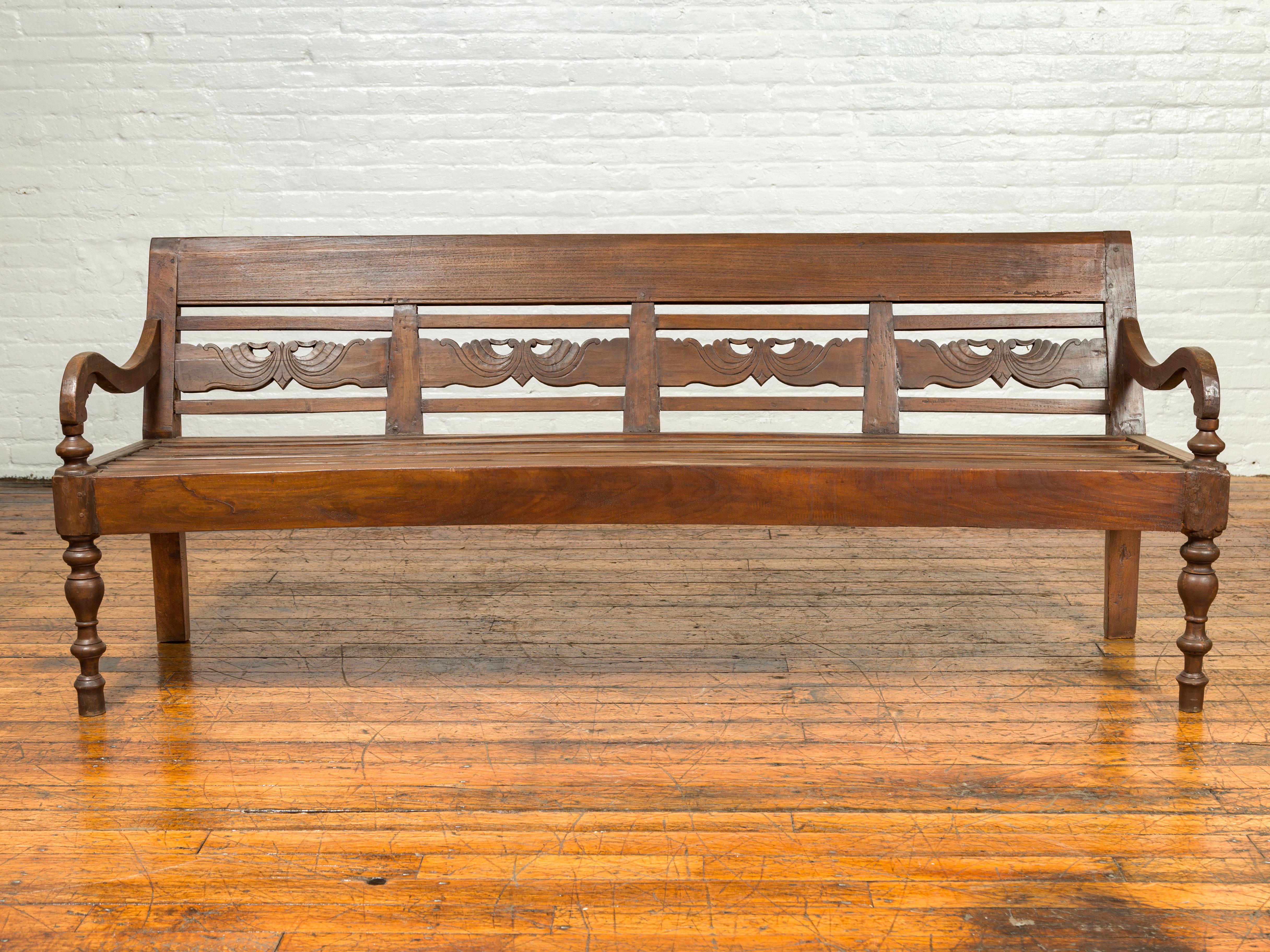 A Dutch Colonial Indonesian wood garden bench from the 19th century, with carved back and scrolling arms. Crafted in Indonesia during the later years of the 19th century, this Dutch Colonial bench features a slanted back carved with scrolling