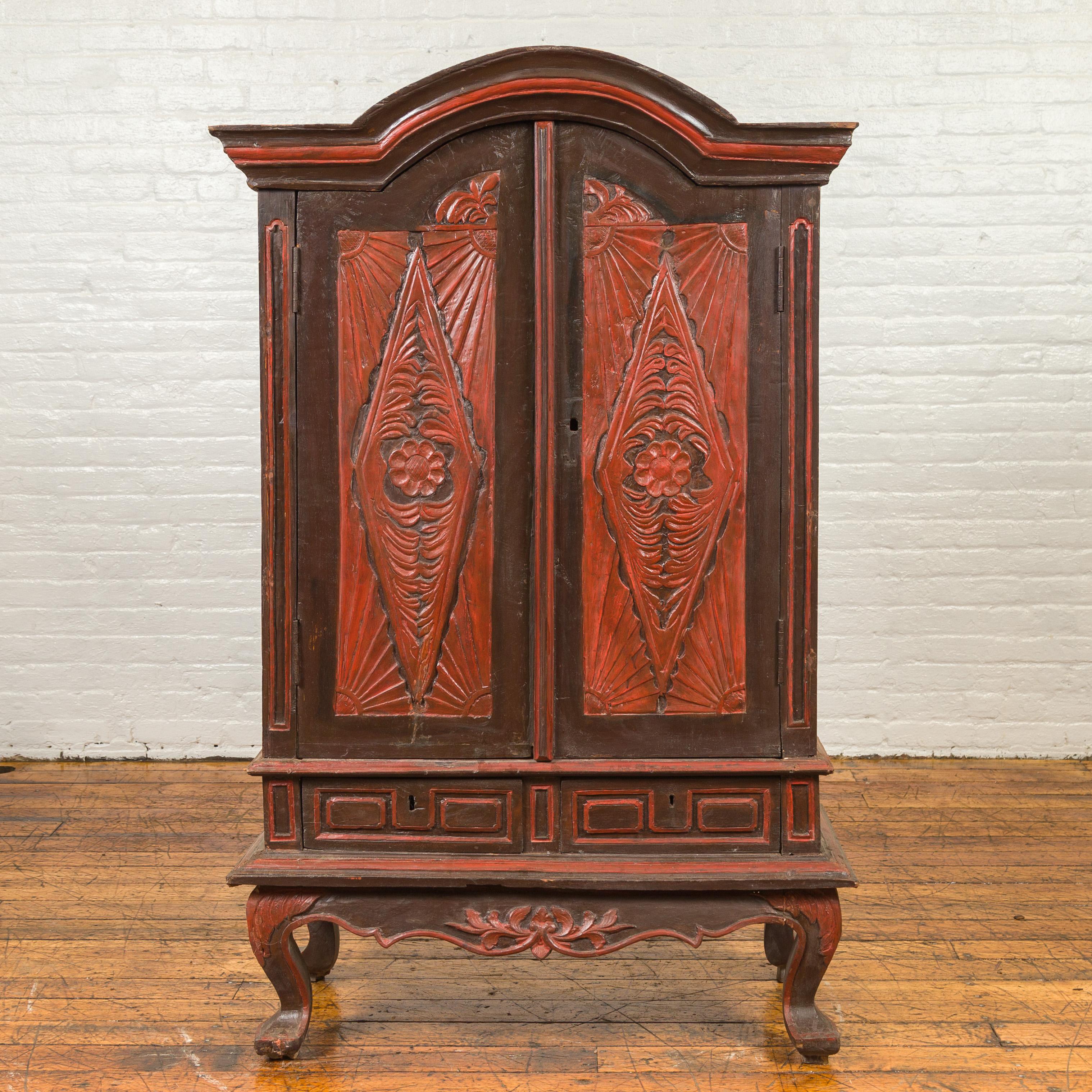 An antique Dutch Colonial cabinet from the late 19th century, with bonnet top, carved panels, cabriole legs and cinnabar / black patina. Crafted in Indonesia during the later years of the 19th century, this cabinet features a bonnet top sitting