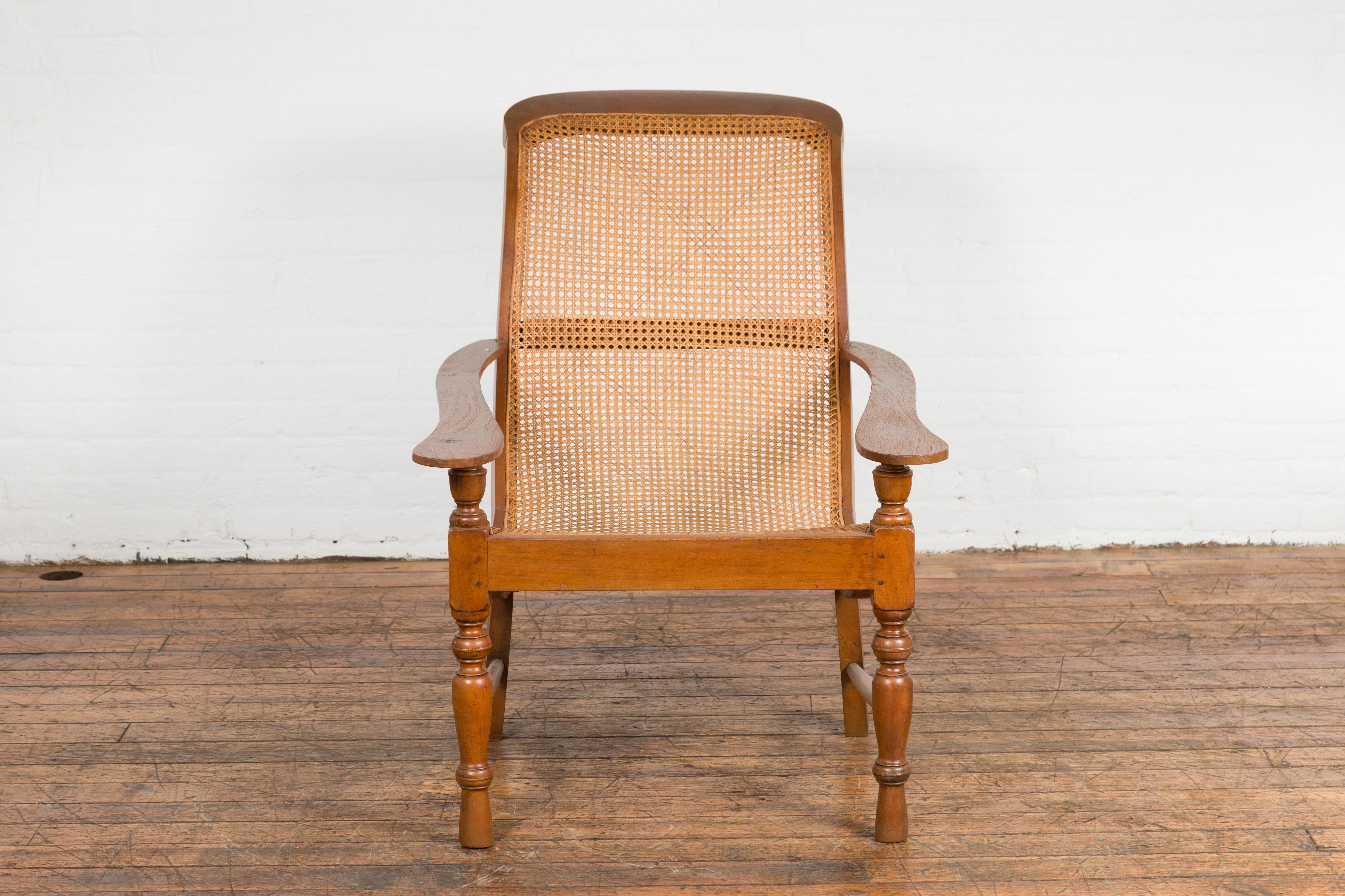 An antique Dutch Colonial Indonesian cane and wood plantation chair from the 20th century with serpentine arms and turned baluster legs. Immerse yourself in the grandeur of Indonesian design with this antique Dutch Colonial plantation chair from the