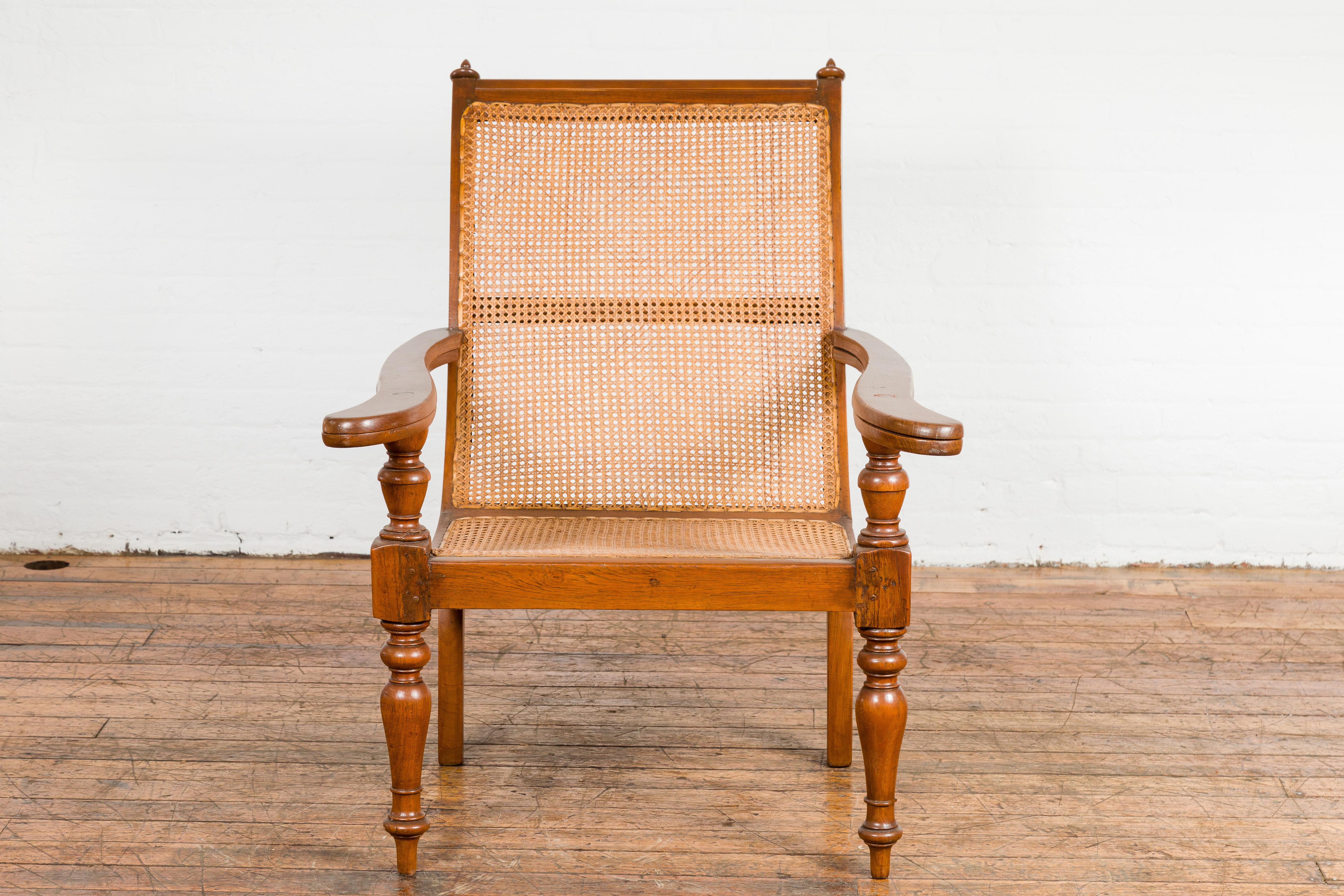 A Dutch Colonial period wood and rattan lounge plantation chair with slanted, curving back, extending arms and petite turned finials. Introducing a stunning piece of history, this Dutch Colonial period wood and rattan lounge plantation chair