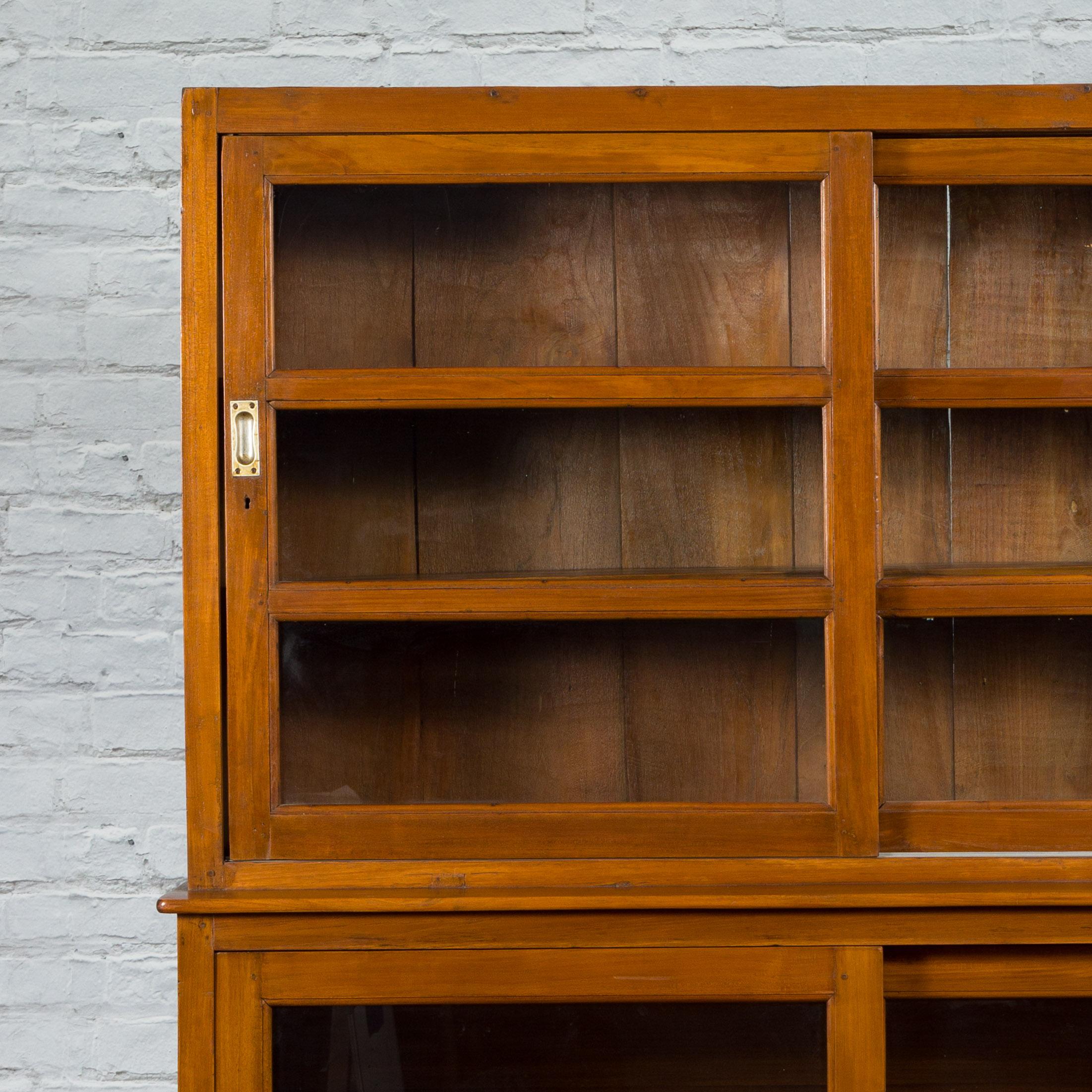 A vintage Indonesian Dutch Colonial style teak wood bookcase from the mid 20th century with sliding glass doors. Created in Indonesia during the Midcentury period, this teak (a very sturdy wood) bookcase features a linear silhouette perfectly