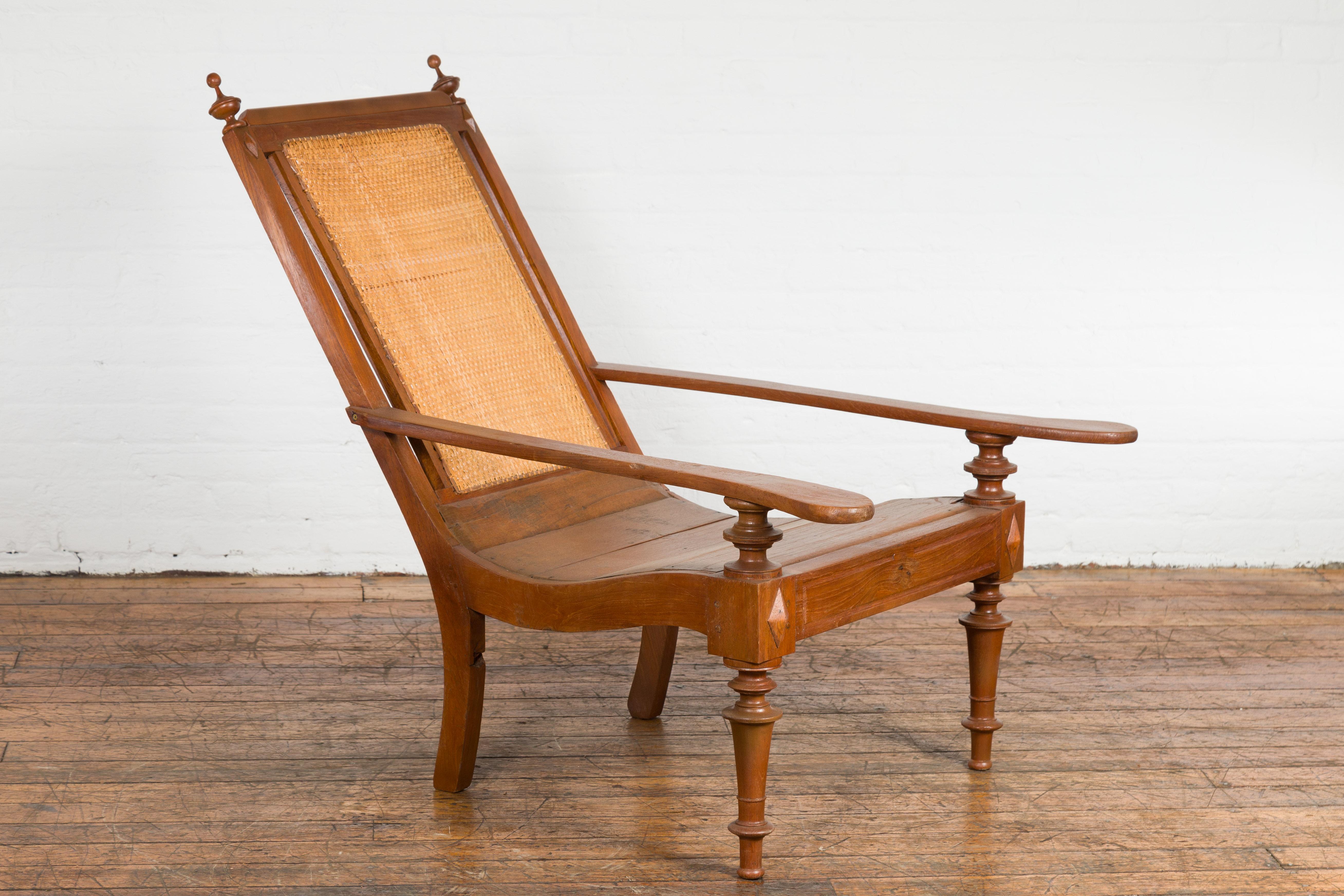 A Dutch Colonial wooden lounge chair from the early 20th century with slanted rattan back, long arms, carved diamond motifs on the knees and turned legs in the front. Nestled in the legacy of the early 20th century, this wooden lounge chair exudes a