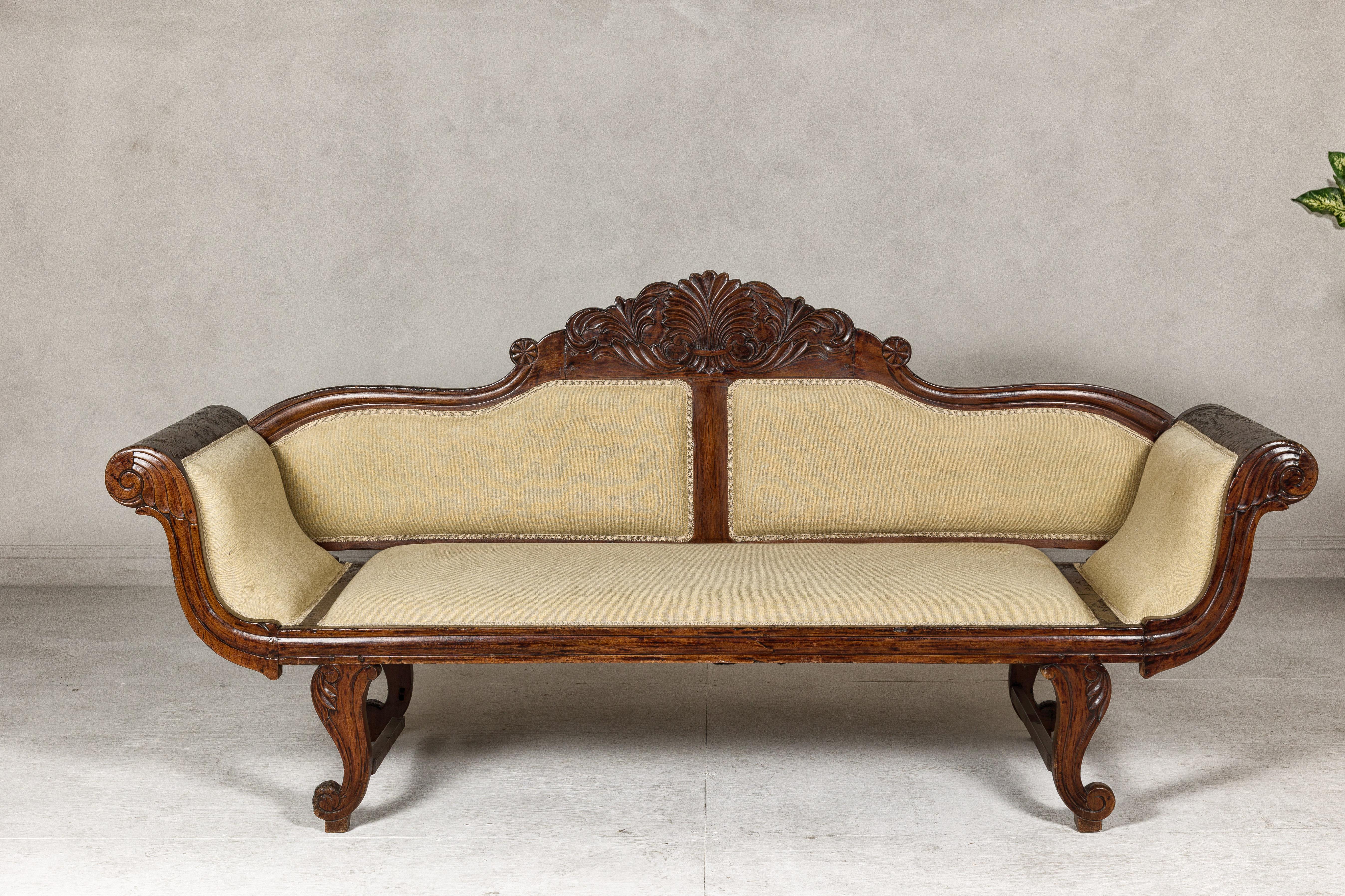 A Dutch Colonial period wooden settee from the early 20th century with carved crest, out-scrolling arms, cabriole legs and used fabric. This early 20th-century Dutch Colonial period wooden settee from South-East Asia is a fine example of classic