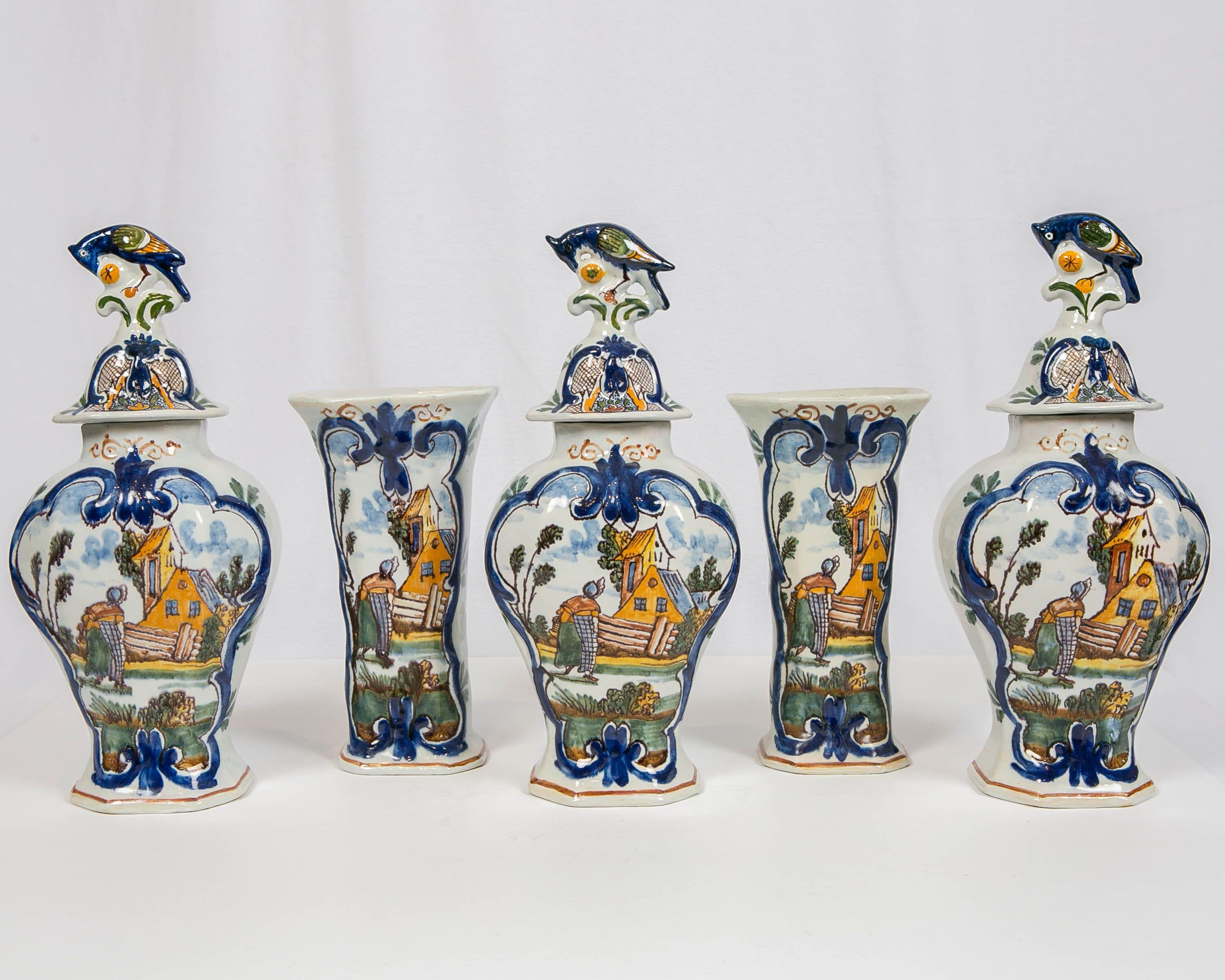 A five-piece mantle garniture comprising three covered vases and a pair of beakers. Made in the Netherlands circa 1750, the jars are each hand-painted on the front with a brightly colored polychrome scene showing a Dutch peasant woman returning home