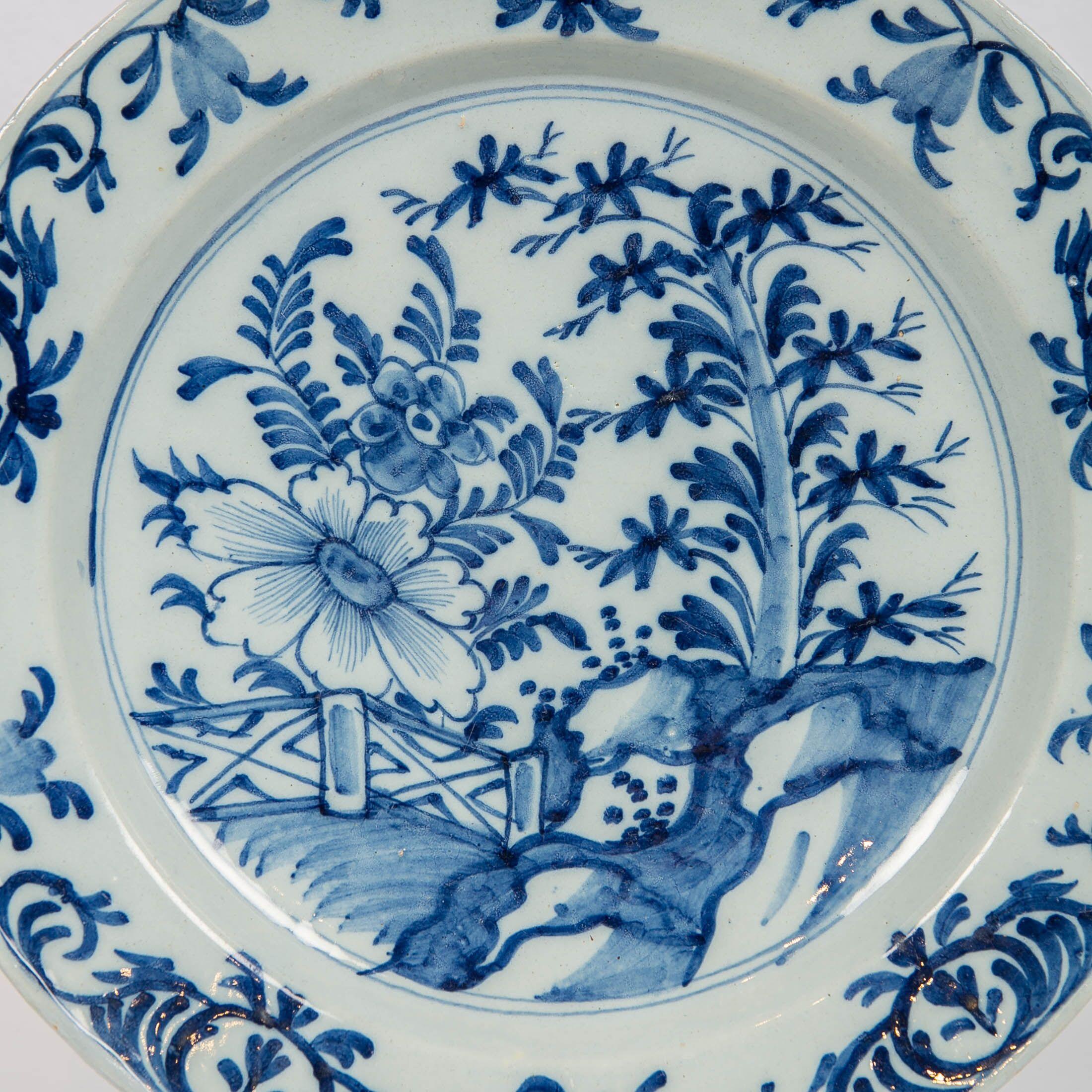 We are pleased to offer this antique Dutch delft blue and white charger made in the 18th century, circa 1780. The center is painted in a variety of shades of cobalt blue showing a garden scene with a pine tree, a large peony, and a garden fence. The