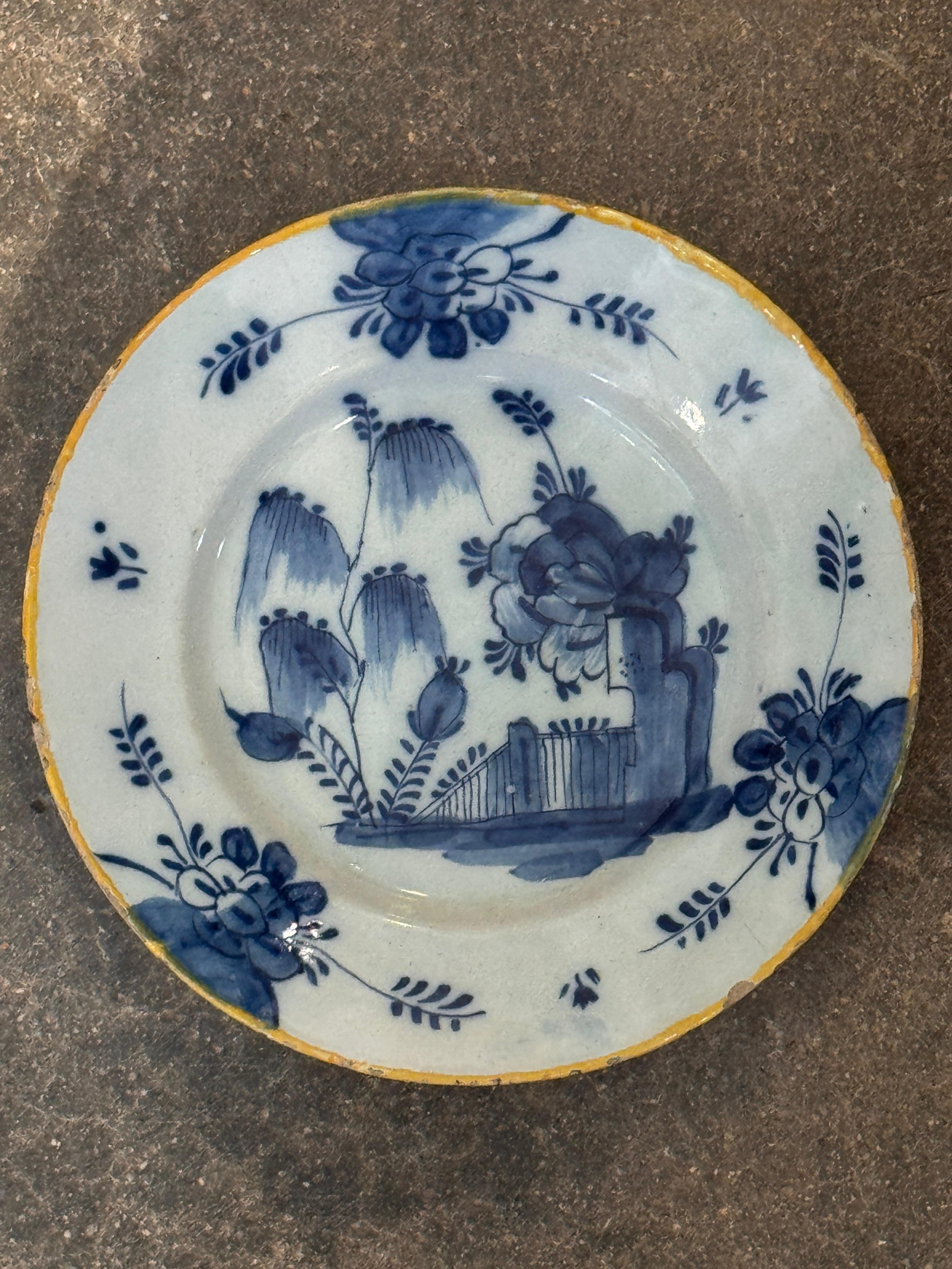 Assiette de Delft du XVIIIe siècle peinte en bleu et blanc avec une scène de jardin floral et une clôture stylisée au centre, encadrée par des gerbes de fleurs éparses. Le bord est rehaussé d'une bordure jaune, un détail que l'on retrouve souvent