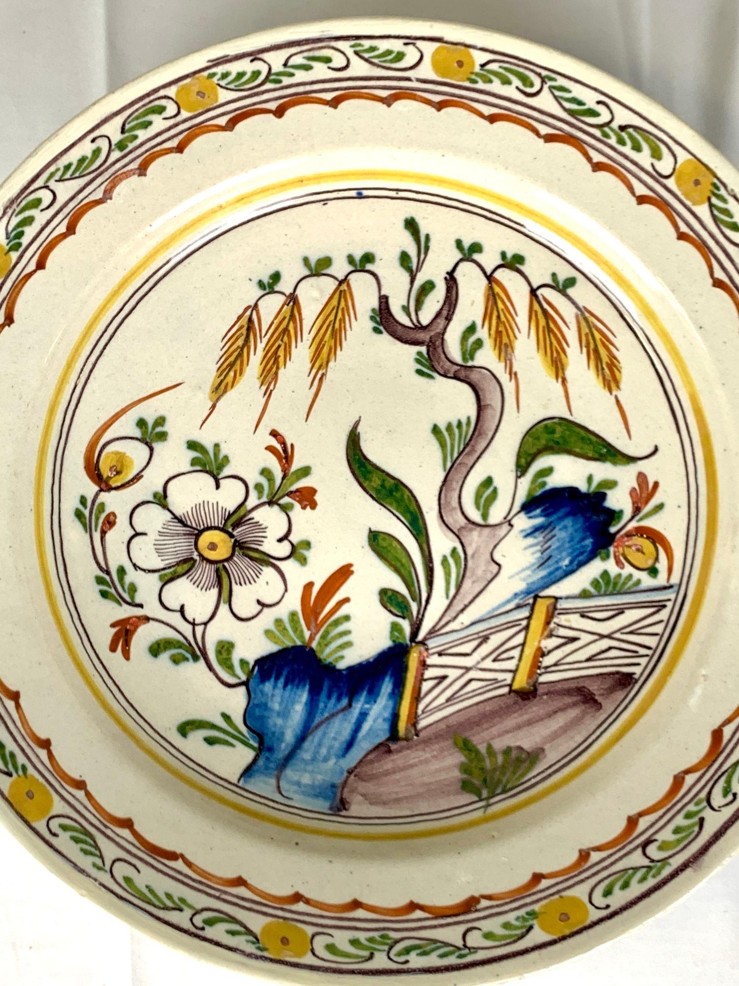 This antique Dutch Delft charger is hand painted in a vibrant array of polychrome hues, including cobalt blue, green, yellow, iron red, ochre, and manganese purple.
We see a beautiful flower-filled garden featuring a willow tree with a purple trunk