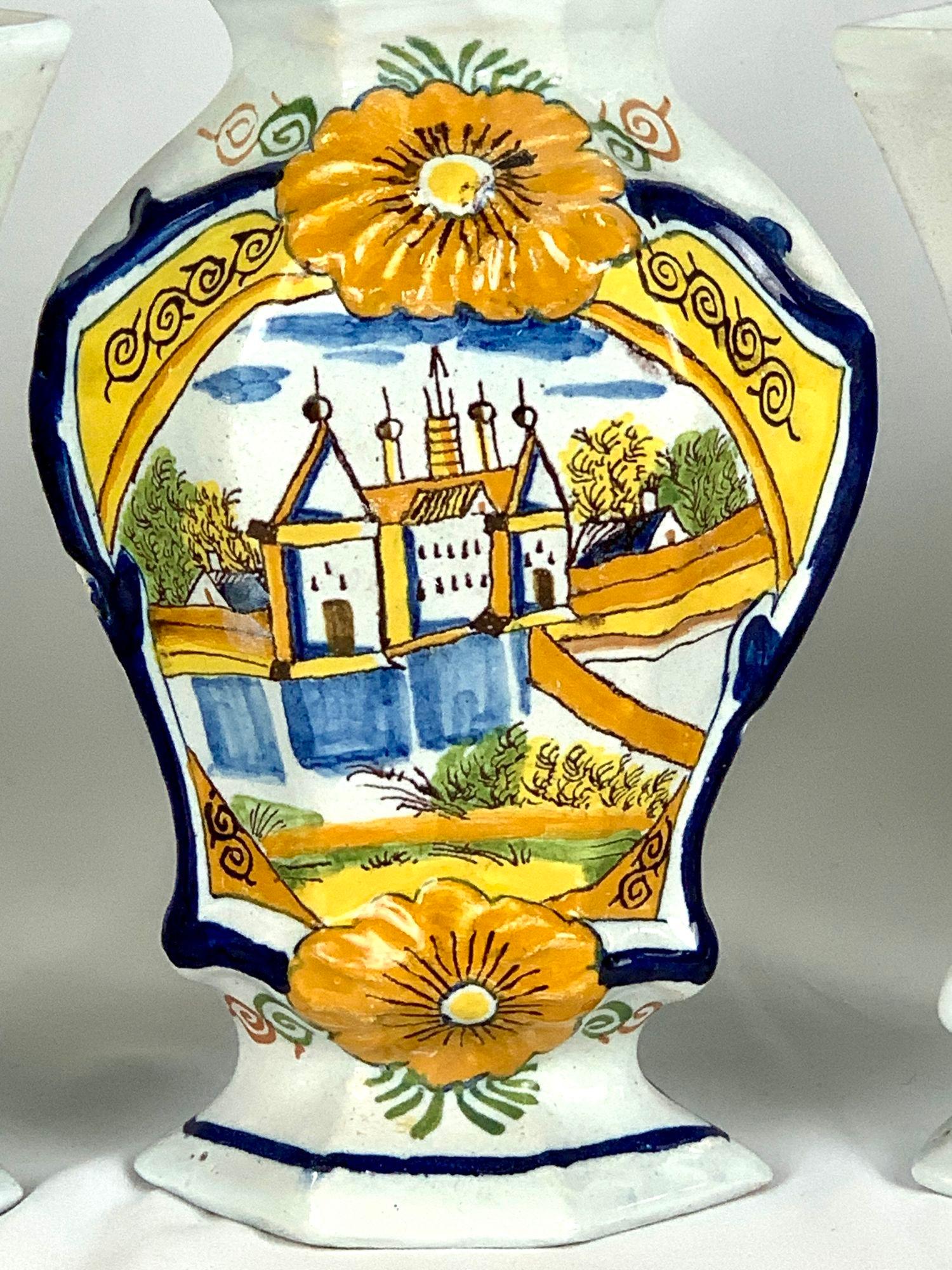 This Dutch Delft mantle garniture comprises five pieces: three lidded vases and a pair of flute-shaped vases.
It was crafted in the Netherlands around 1780.
The design features a hand-painted, naive scene of a castle behind a moat filled with blue