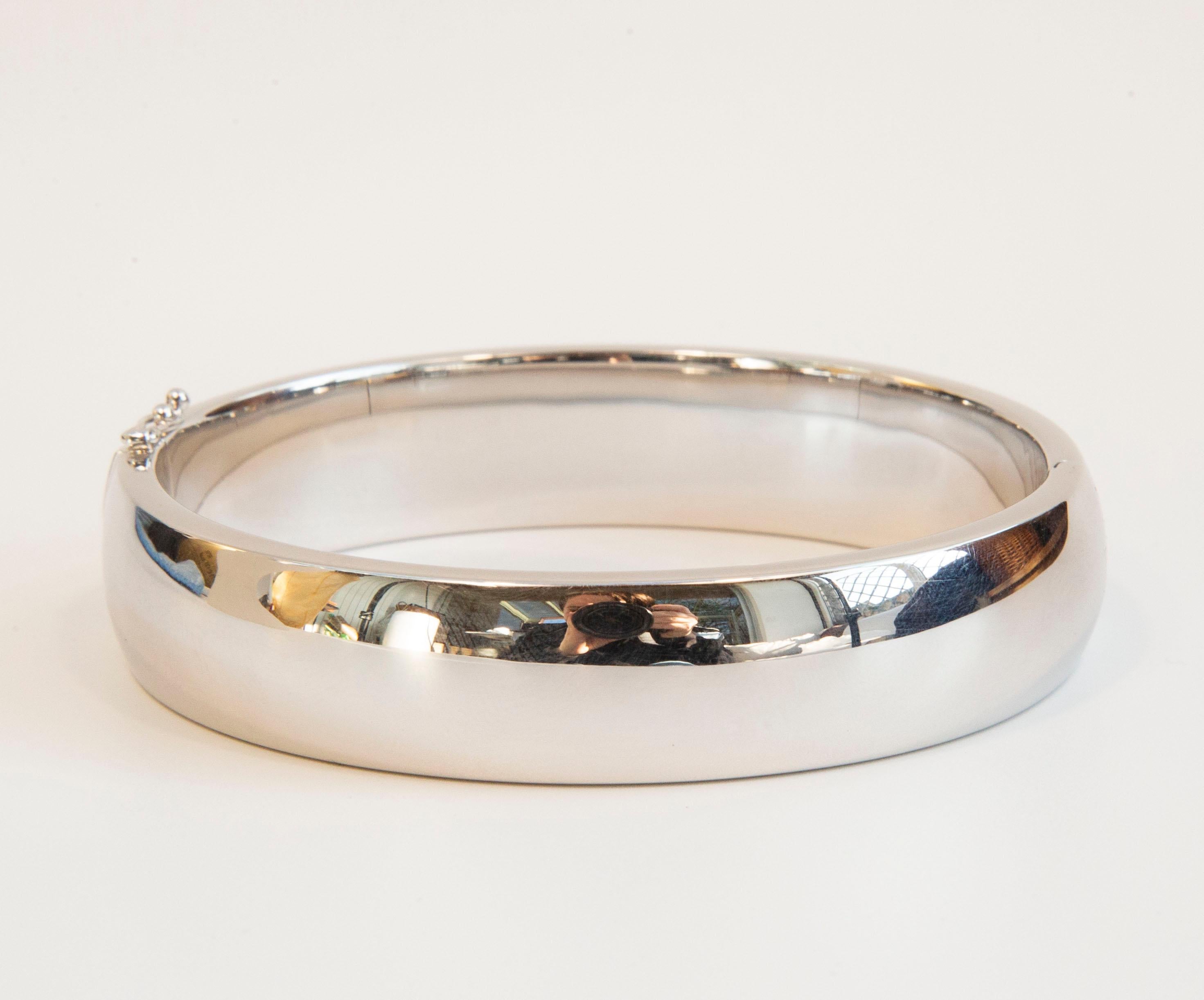 Un bracciale bangle moderno e minimalista in argento sterling. Il bracciale presenta una finitura lucida e liscia. Si chiude con una cerniera e una chiusura a pressione e, per maggiore sicurezza, con un lucchetto di sicurezza a forma di otto, che