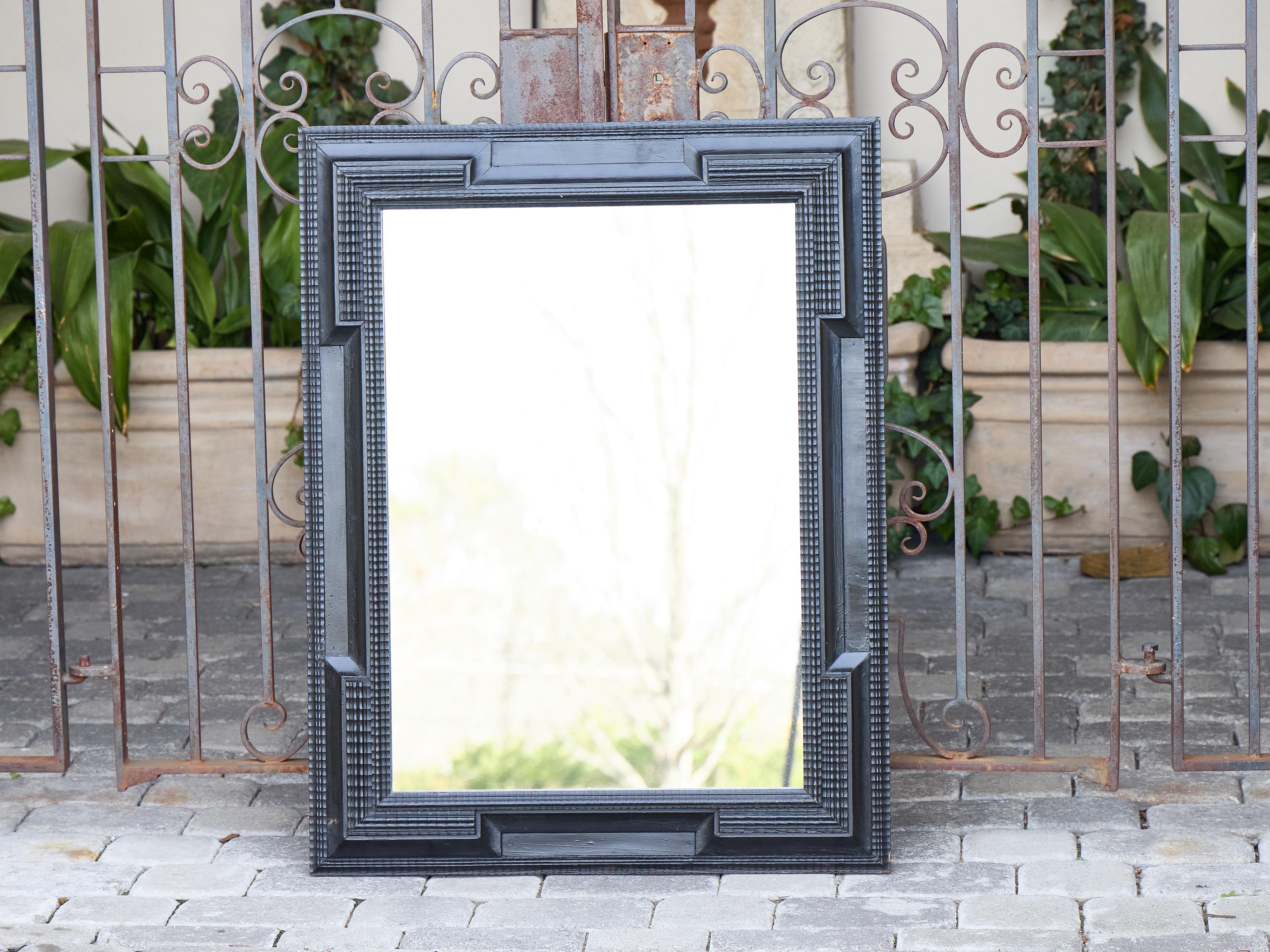 This Dutch Baroque Revival mirror from circa 1900 features a bold ebonized frame with stepped, architectural lines and crisp corner detailing that draws from 17th-century Dutch design traditions. The deeply recessed molding and rhythmic grid-carved