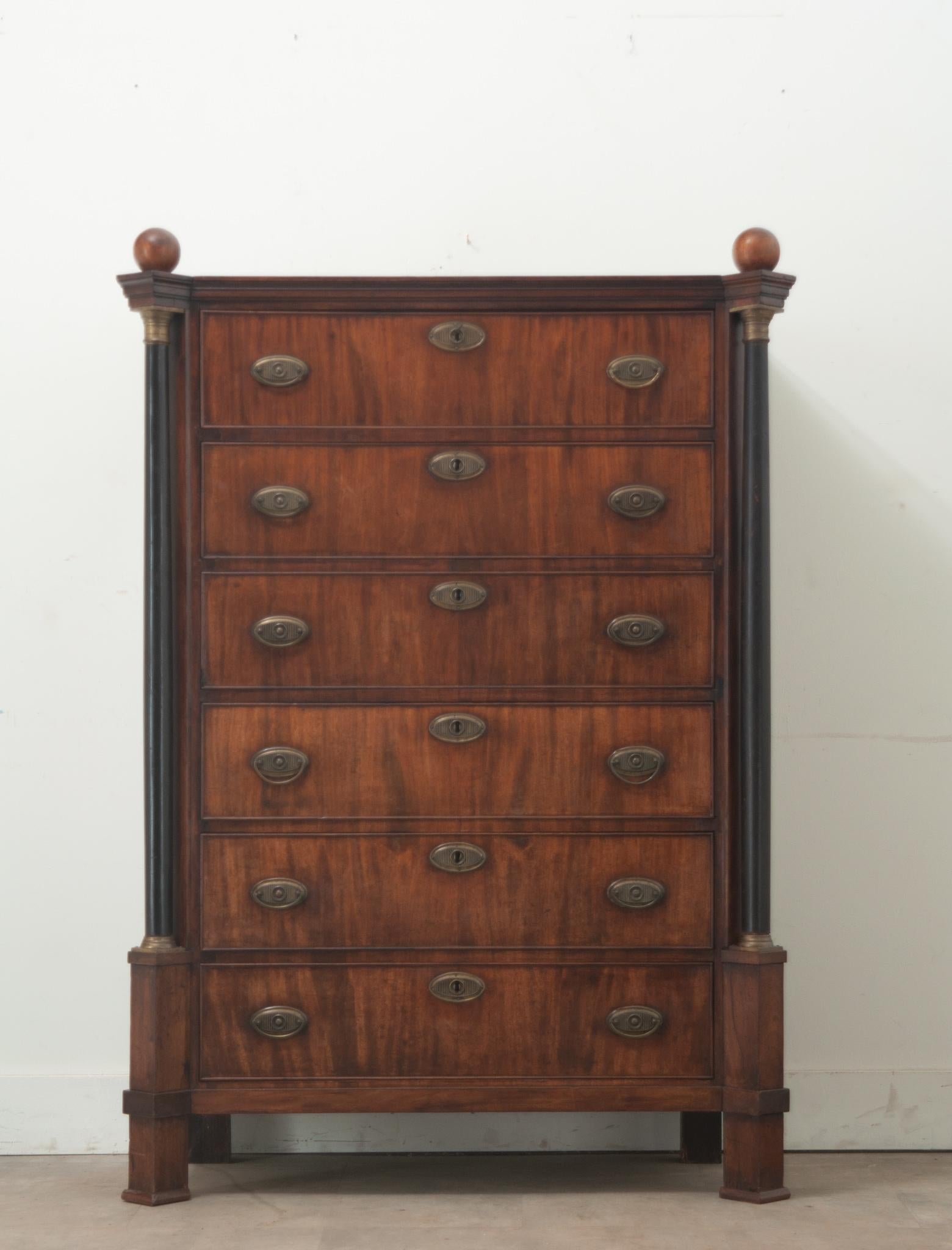 A tall Dutch Empire style chest made of mahogany and ebonized wood during the 1800’s. This mahogany case piece has six bookmatched drawer fronts each with two ring drawer pulls for easy accessibility. Ebonized columns topped with ball finials flank