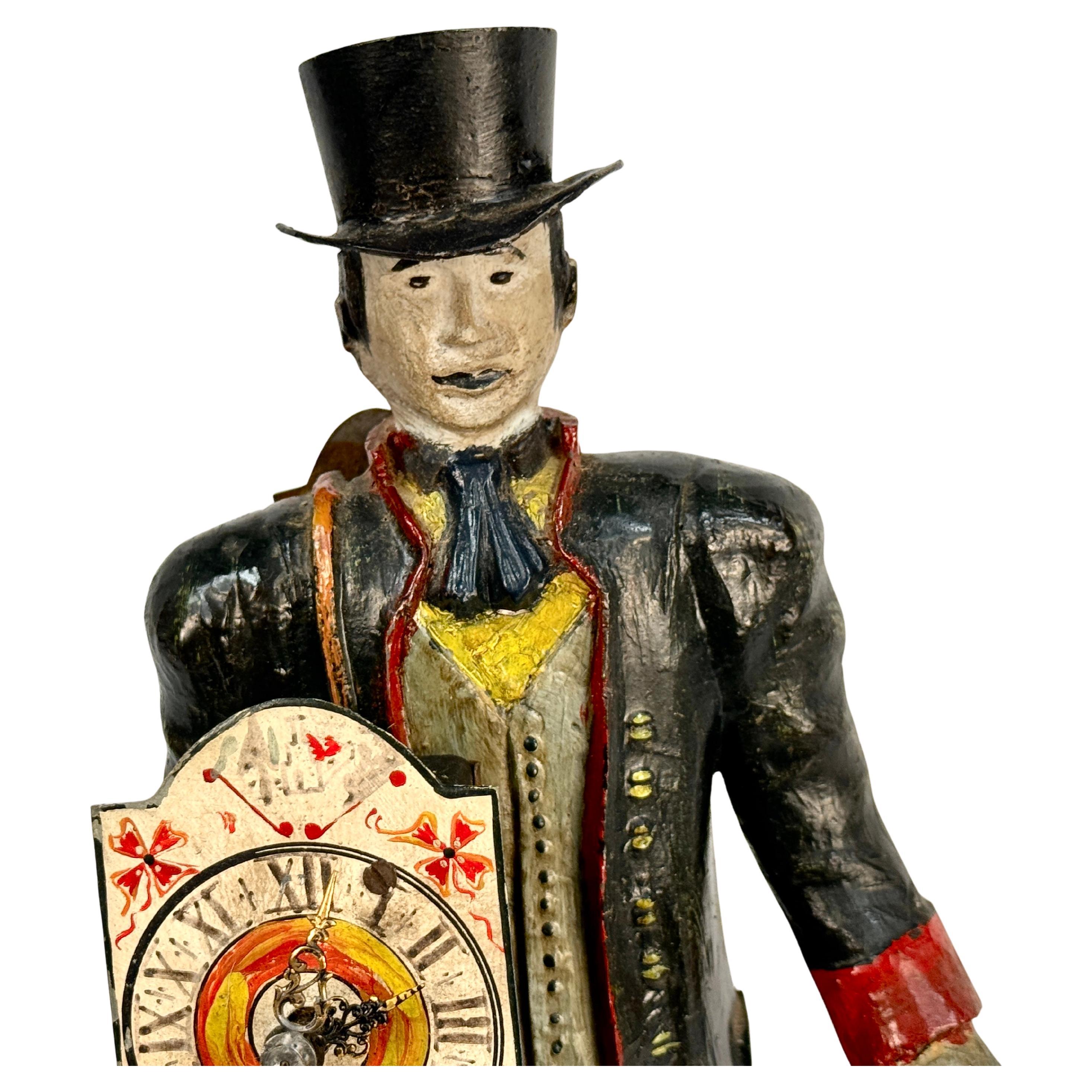 Cette horloge de colporteur hollandaise figurative du XIXe siècle est caractérisée par un colporteur masculin vêtu d'un pardessus noir et d'un chapeau haut de forme, tenant une horloge dans sa main droite. Ses vêtements sont ornés de manchettes