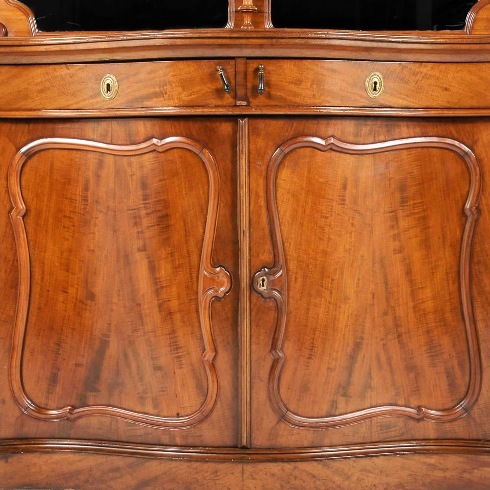 A Continental European serpentine-front mahogany server, or buffet, with an elaborately shaped and carved mirror-back over a two-door cabinet with upper single drawer, circa 1870.

     