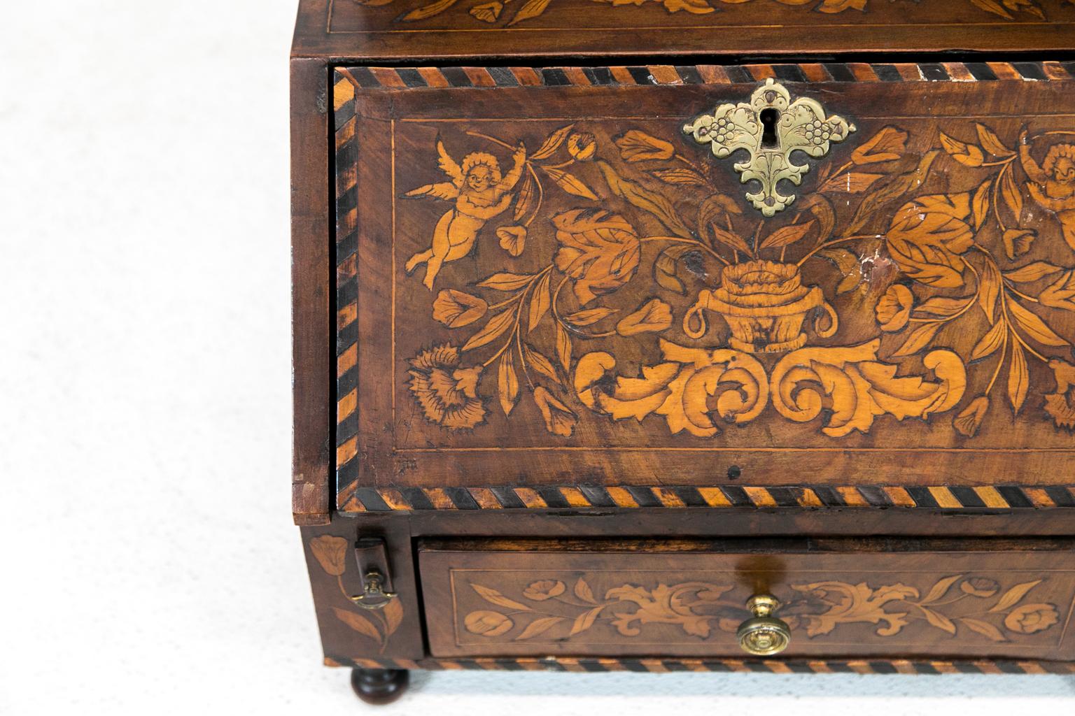 Dutch Marquetry miniature slant top desk has a mirror and is inlaid with cupids, flowers, and baskets and has carved mask finials. It has the original brass hardware.