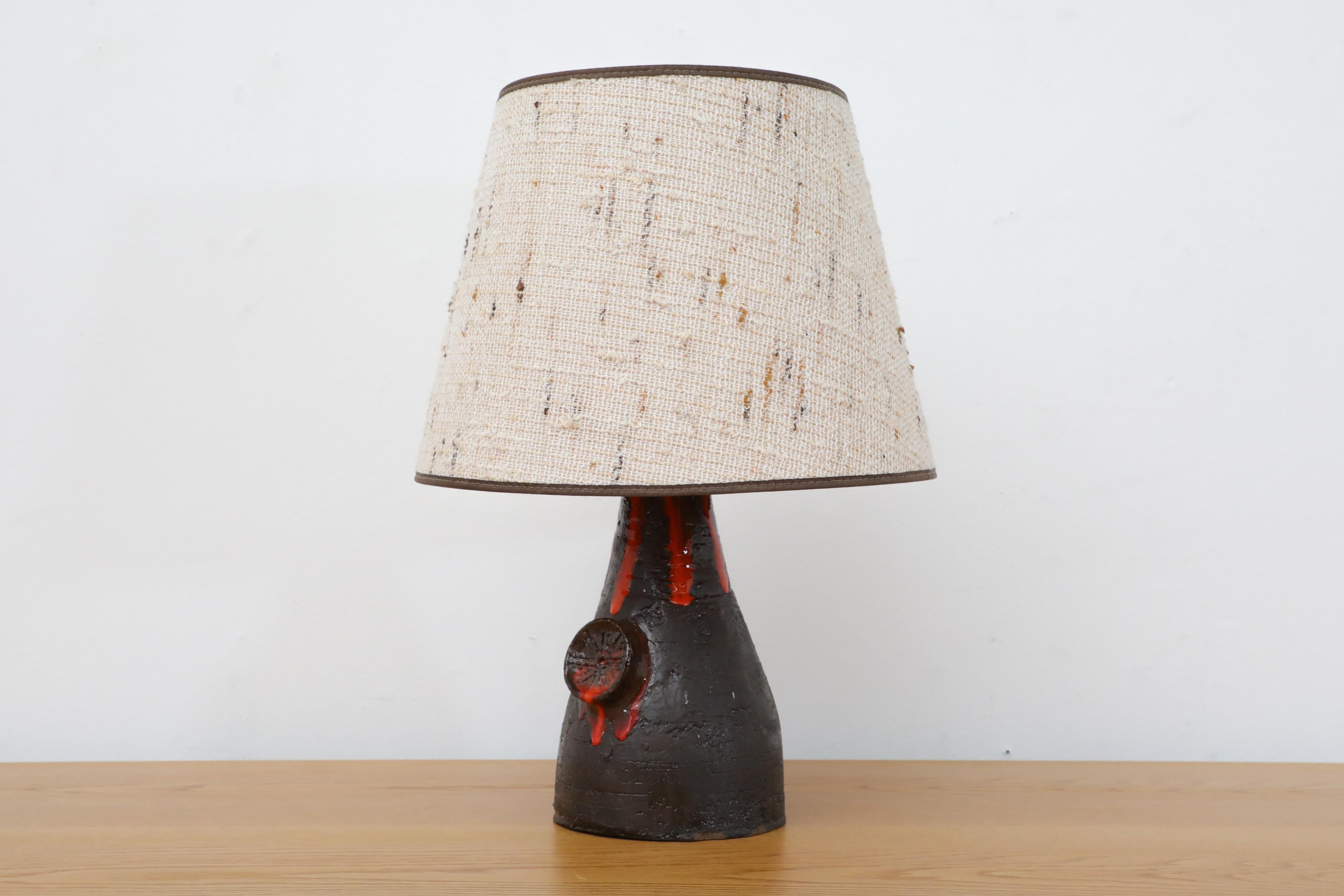 Lampe de table hollandaise du milieu du siècle en céramique émaillée noire avec une impression volcanique et une glaçure rouge ressemblant à du feu. L'abat-jour est un tambour conique en tissu. En état d'origine avec une usure conforme à l'âge et à